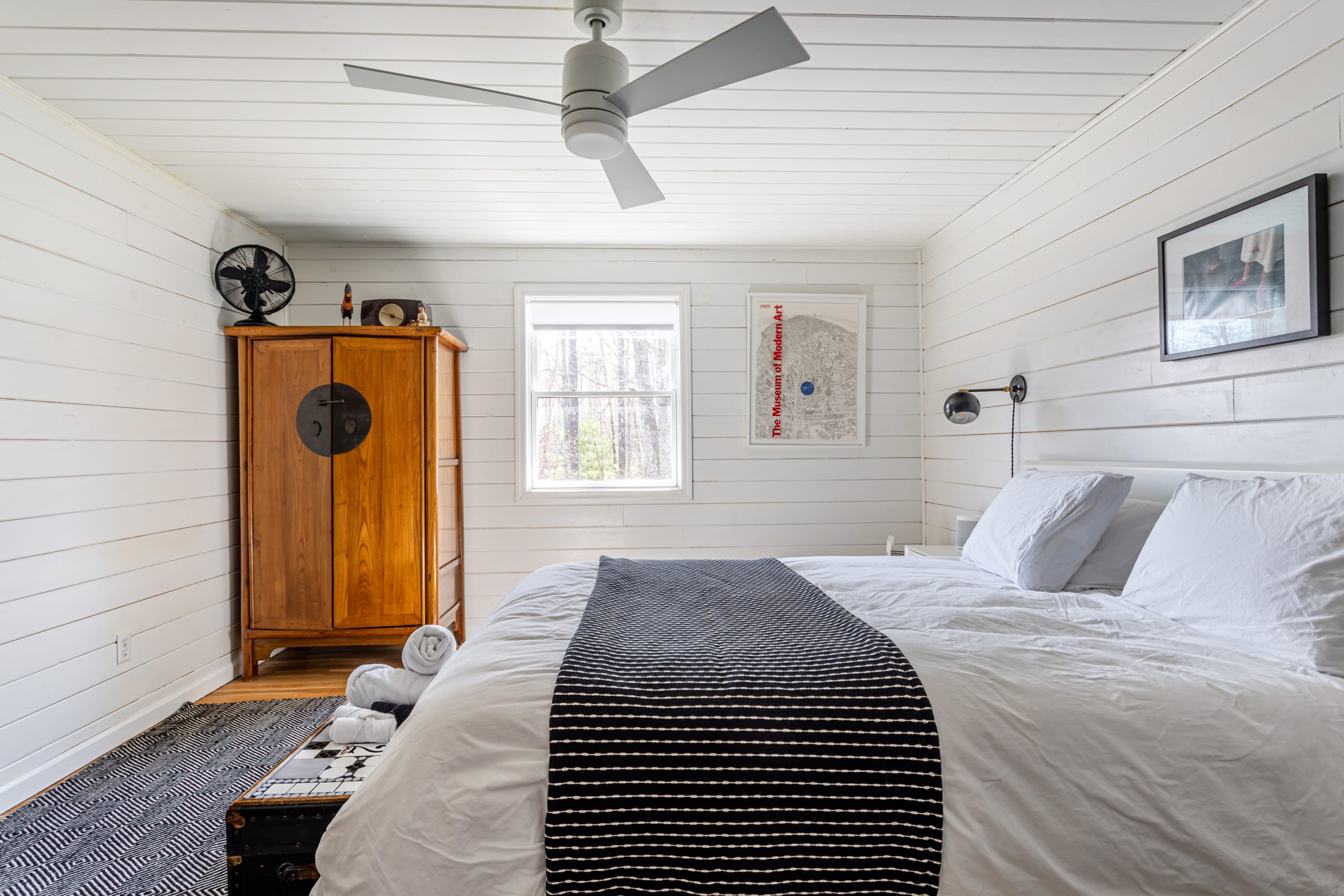 Another view of the bright bedroom, showcasing white shiplap walls and ceiling, a white bed with striped throw, a large wooden armoire, and a window offering a glimpse of the outdoors.
