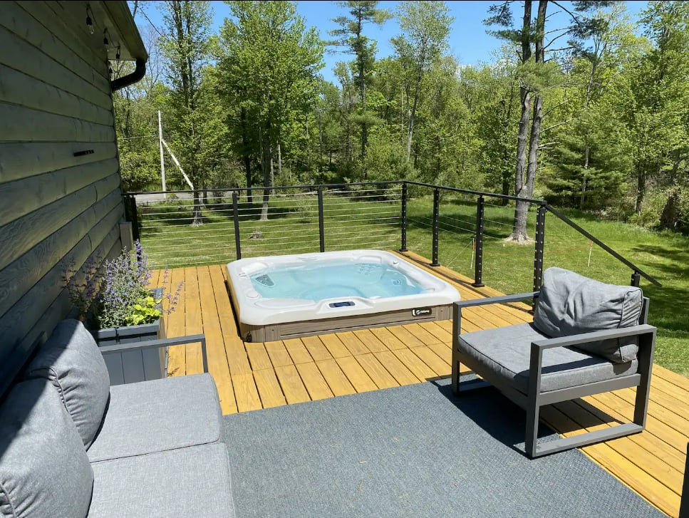 A white hot tub is centered on a golden-toned wooden deck, surrounded by modern gray outdoor seating and a dark gray rug. A cable railing offers views of lush green trees and a grassy lawn.