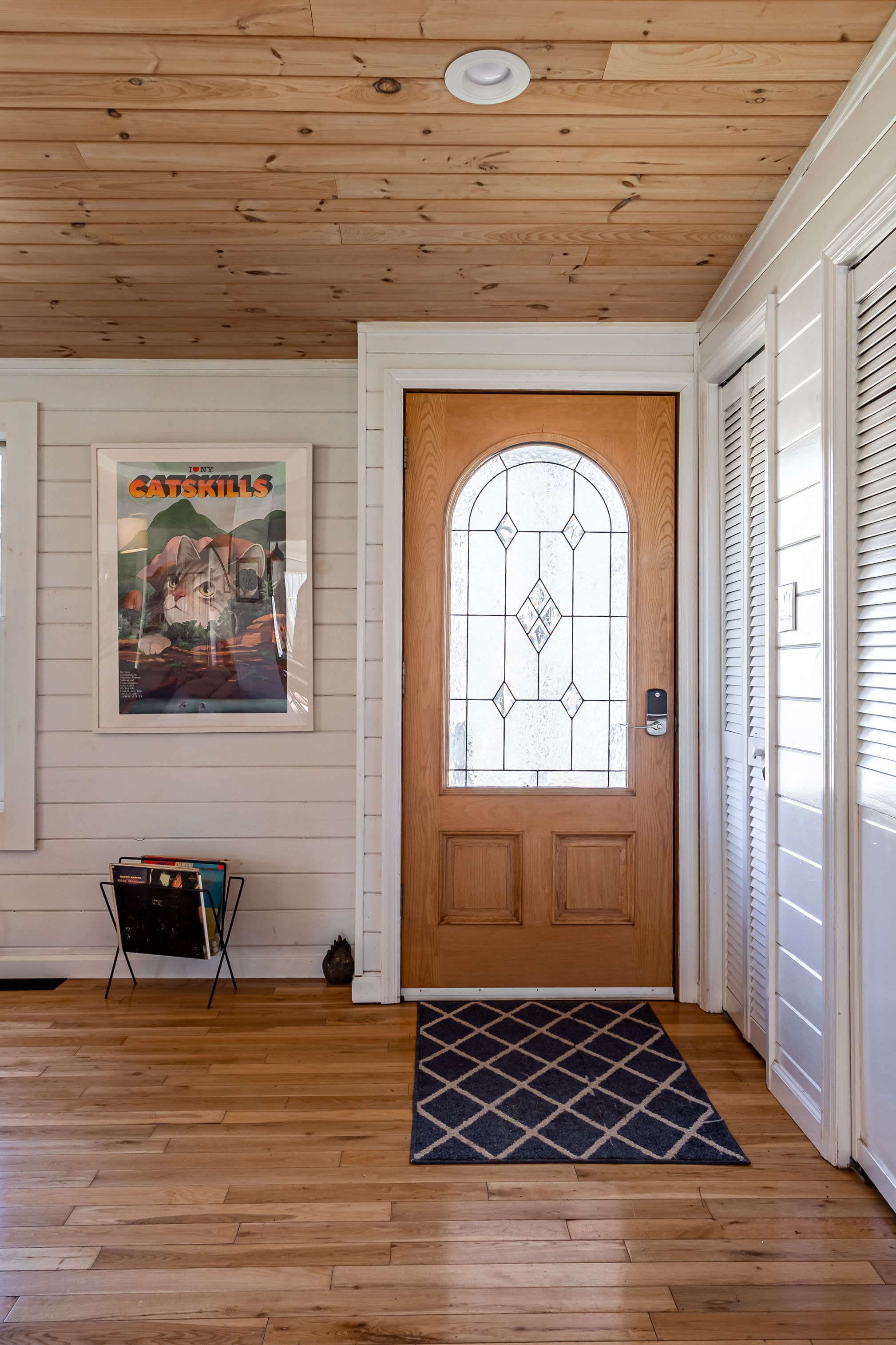 The entryway features a light wood front door with decorative glass, flanked by white shiplap walls and a closet. A "I Love Catskills" poster hangs on the wall, and a small rug is placed at the entrance.