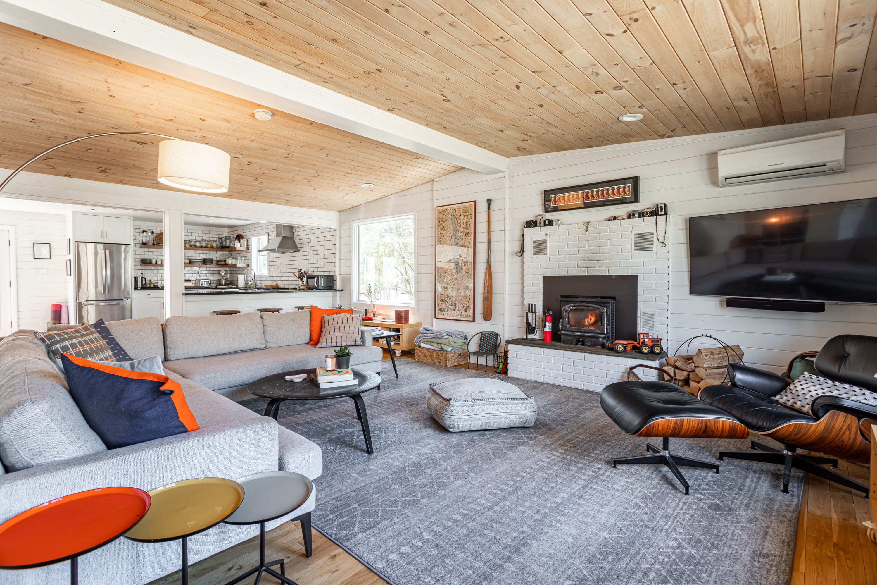 A cozy living room with a large L-shaped sectional sofa, a black coffee table, and a modern Eames lounge chair. The room features white shiplap walls, a light wood plank ceiling, and a white brick fireplace with a wood-burning insert.