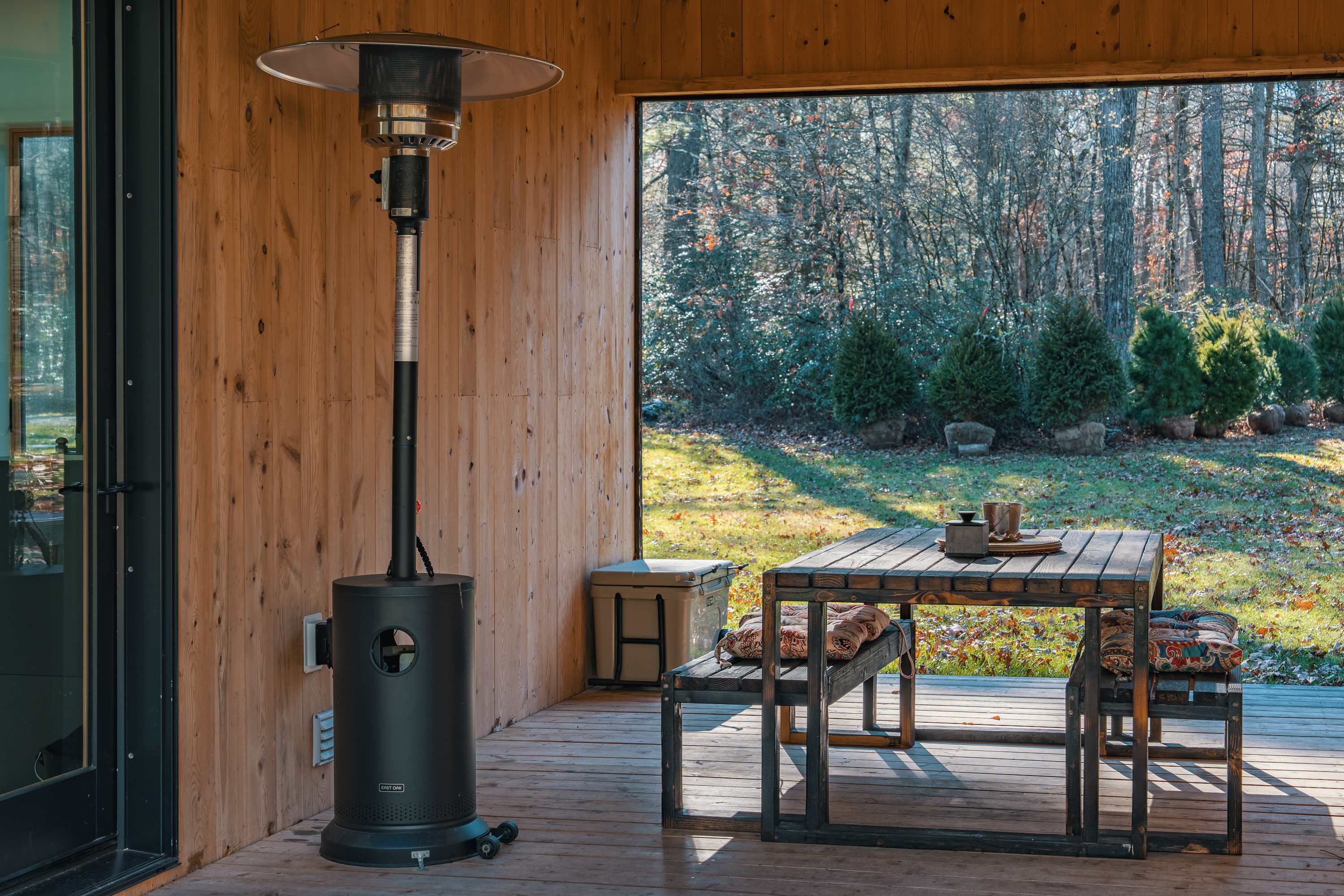 Covered outdoor deck area with wooden flooring and walls, featuring a dark metal patio heater, a rustic wooden dining table with benches, and a view of a wooded backyard.