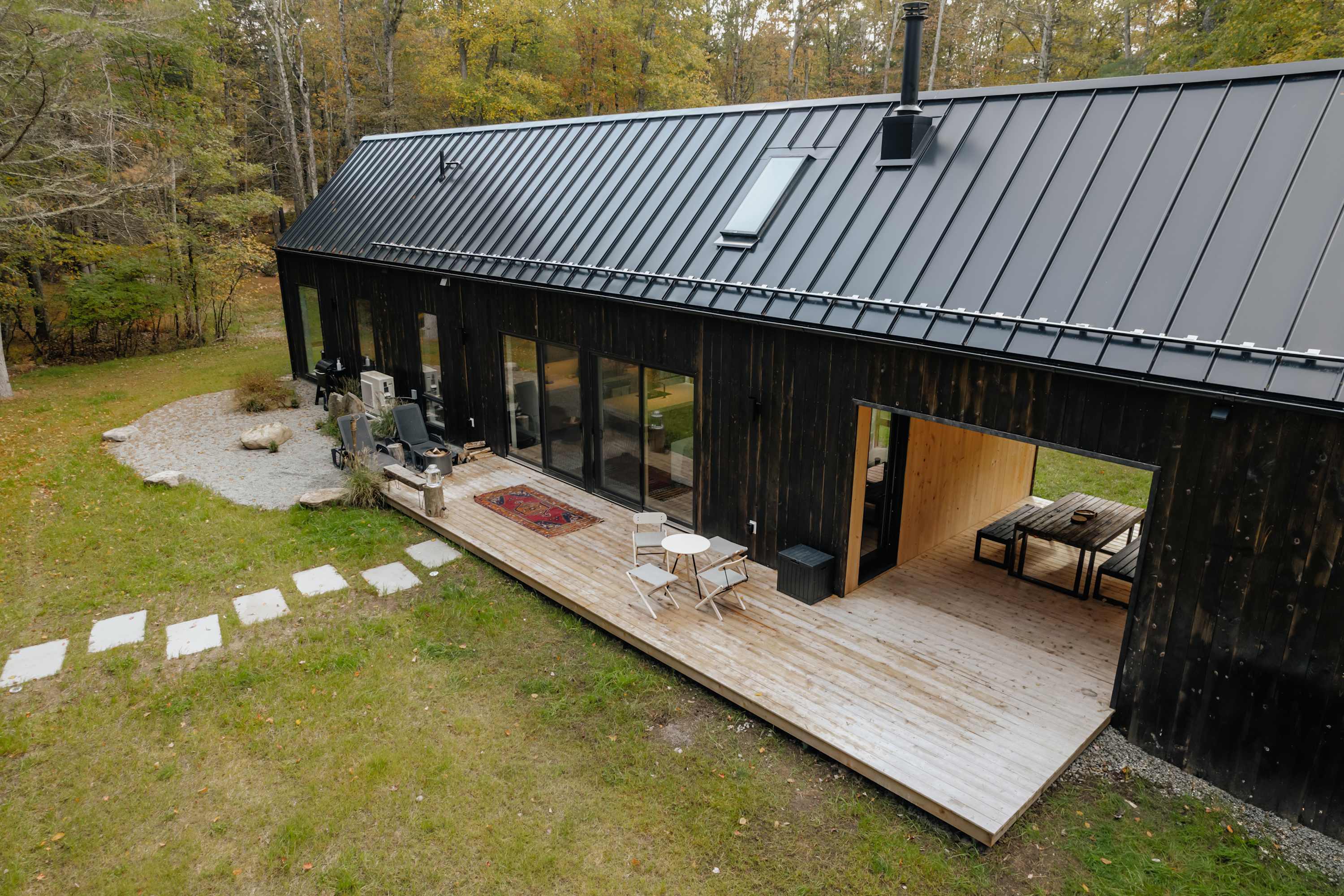 Aerial view of a modern black-clad house with a dark metal roof, surrounded by a lush green lawn and mature trees, featuring a large wooden deck with outdoor seating and a pathway.