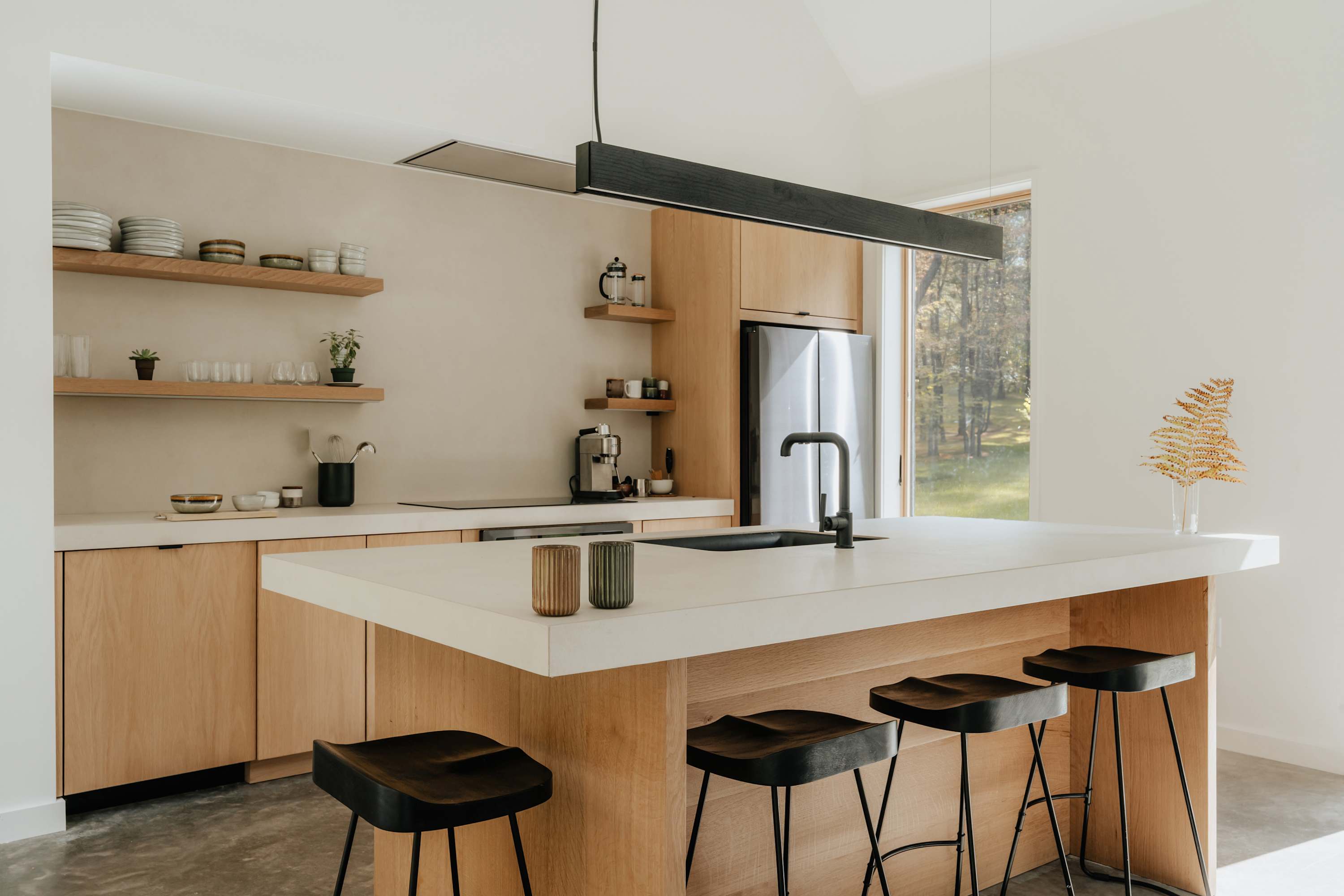 A spacious, minimalist kitchen features light wood cabinetry, white countertops, and a large island with three black bar stools. A long black linear pendant light hangs above the island, complementing the modern aesthetic and open shelving.