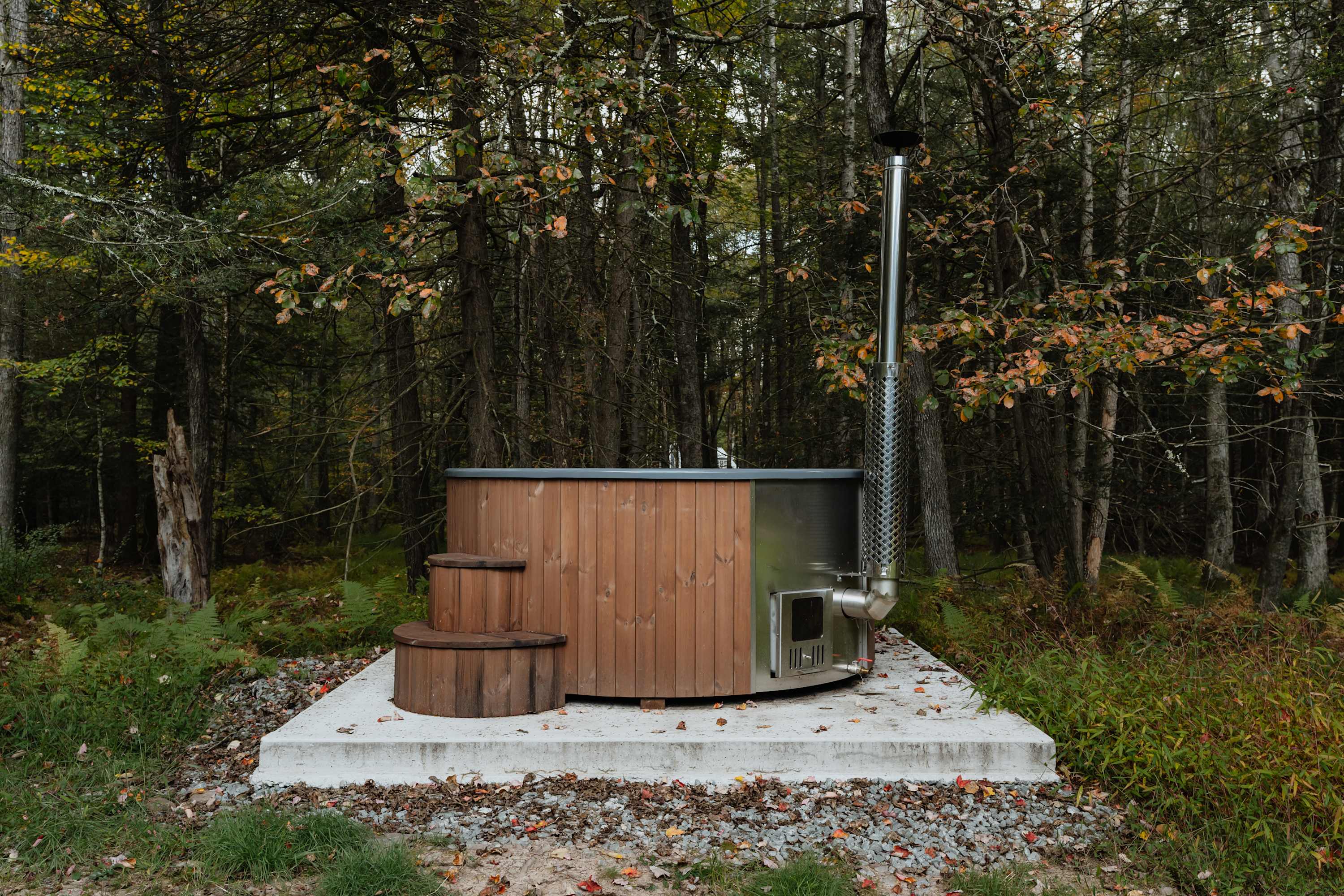 Wood-fired hot tub with a wooden exterior and stainless steel interior, featuring a tall chimney, situated on a concrete pad in a dense autumn forest.