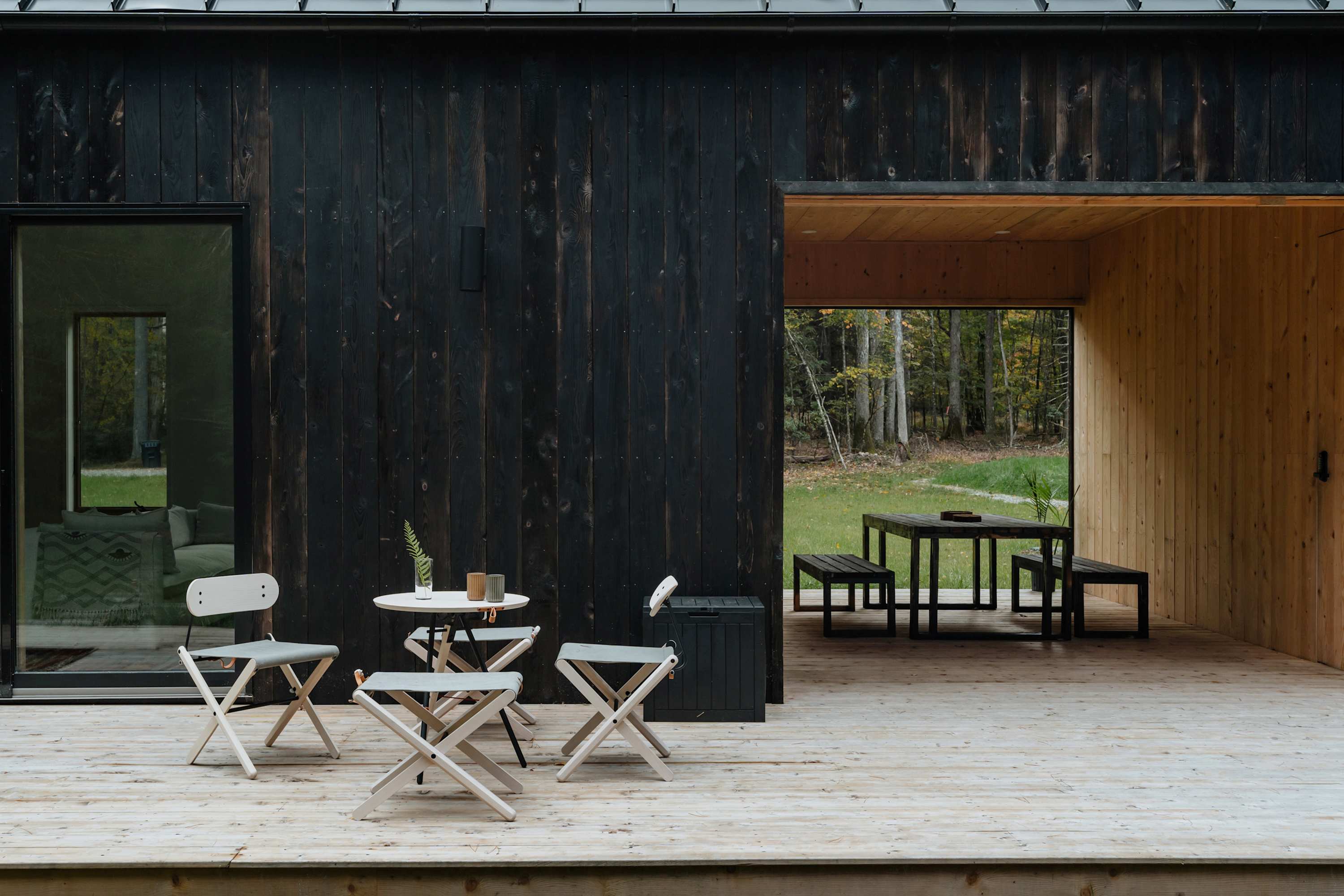 A light wooden deck featuring modern folding chairs and a small table, with a large open-air dining area visible through a wide opening in the dark-clad house. The surrounding forest is visible in the background, offering a serene outdoor living space.