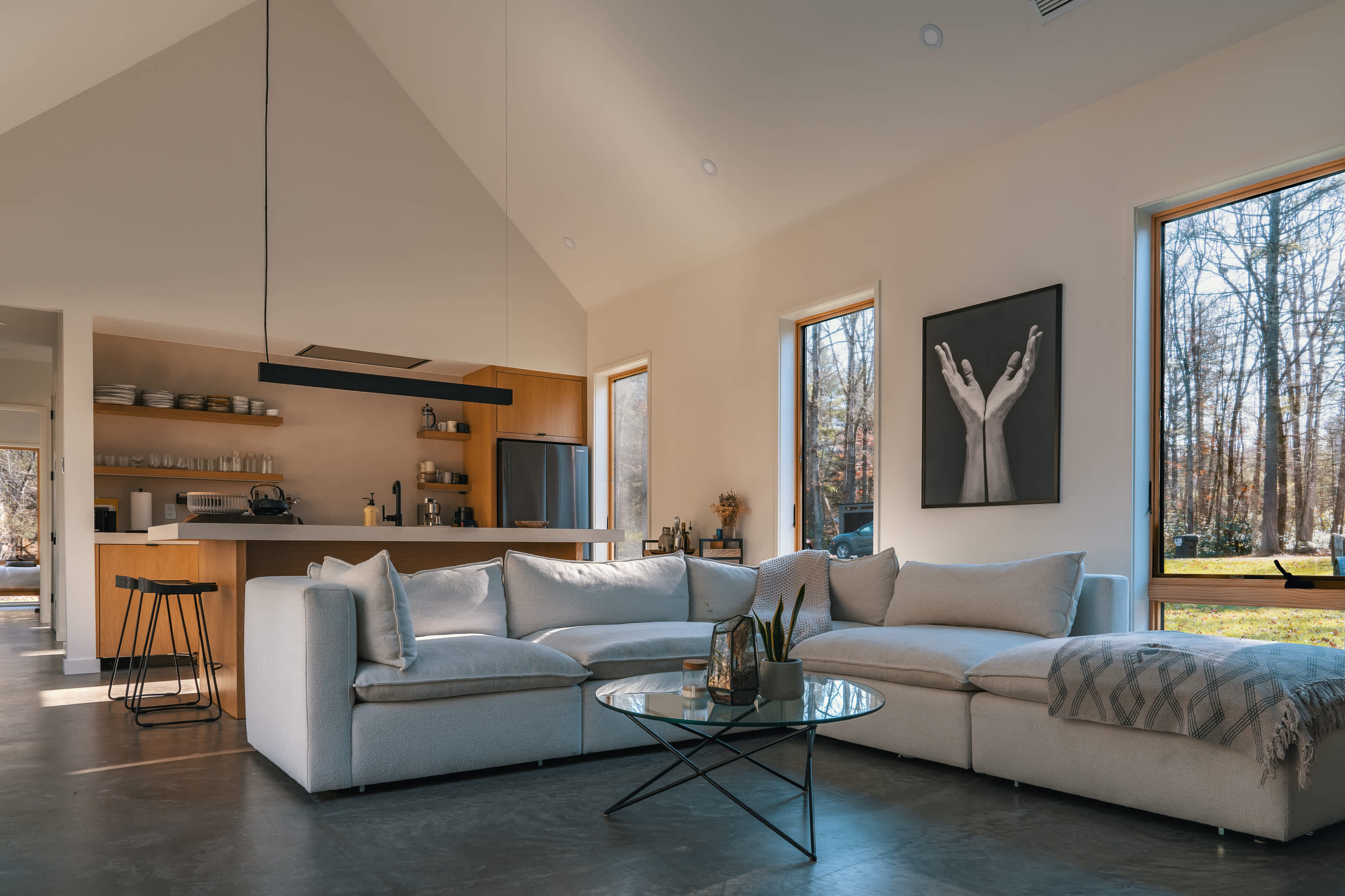 An expansive open-concept living area with a large light-colored sectional sofa and a glass coffee table. The space flows into a modern kitchen with light wood cabinetry, all illuminated by natural light from large windows overlooking a forest.