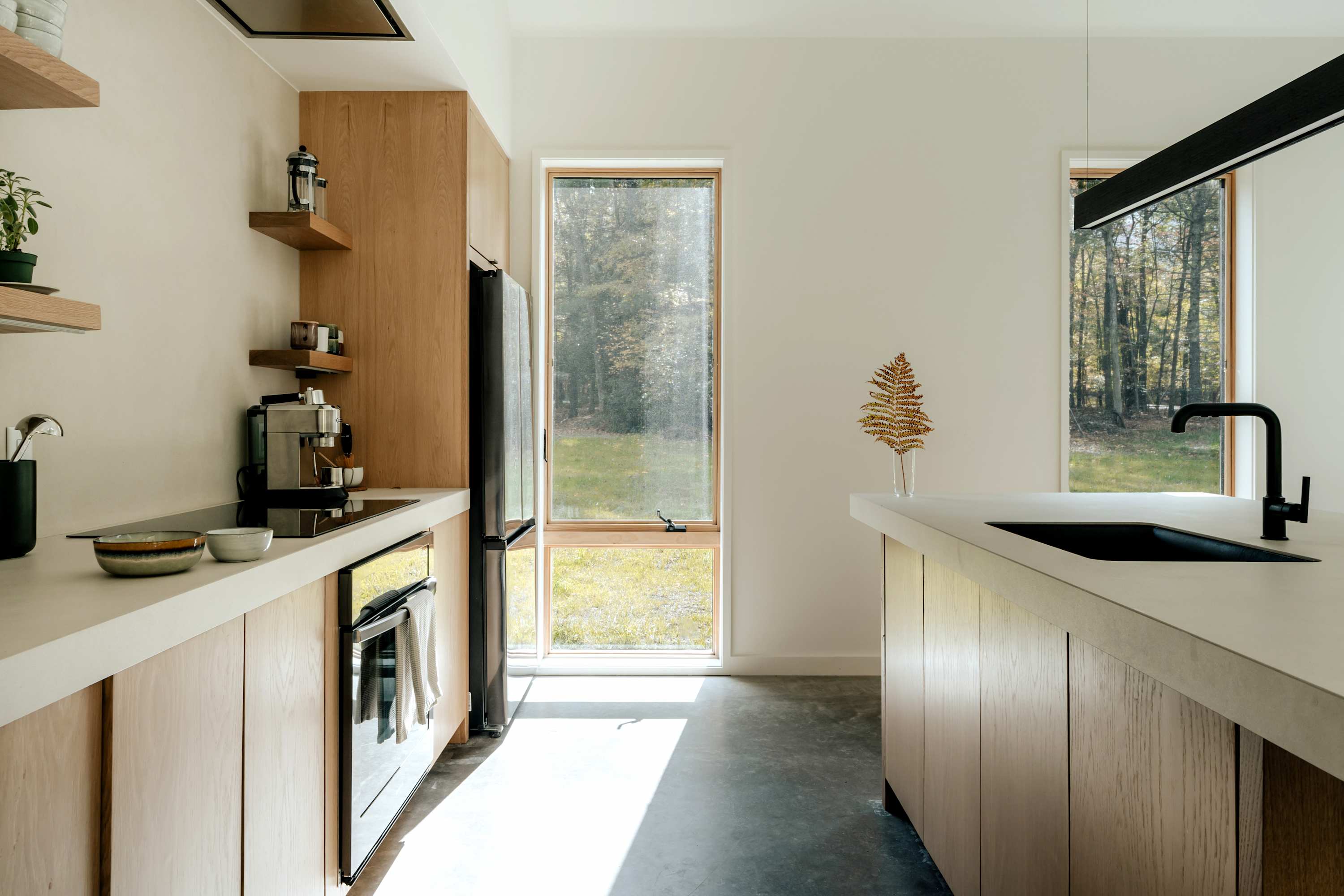A modern kitchen with light wood flat-panel cabinetry, white countertops, and a black built-in oven and refrigerator. Large windows provide ample natural light and views of the outdoors, highlighting the minimalist design.