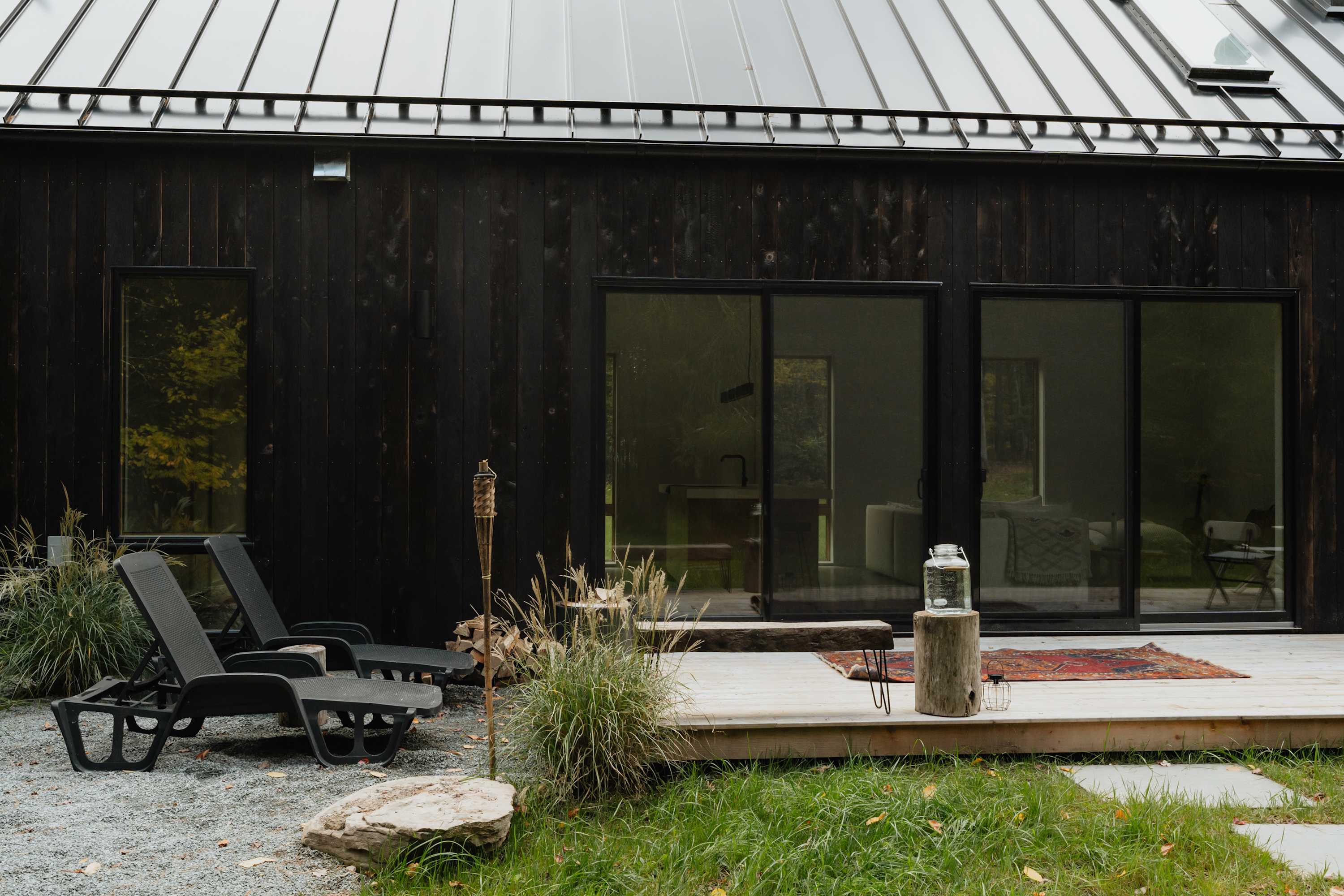 A wooden deck with two black lounge chairs and a small side table made from a tree stump, overlooking a grassy area and surrounded by tall grasses and trees. Large sliding glass doors lead into the house, blurring the lines between indoor and outdoor living.