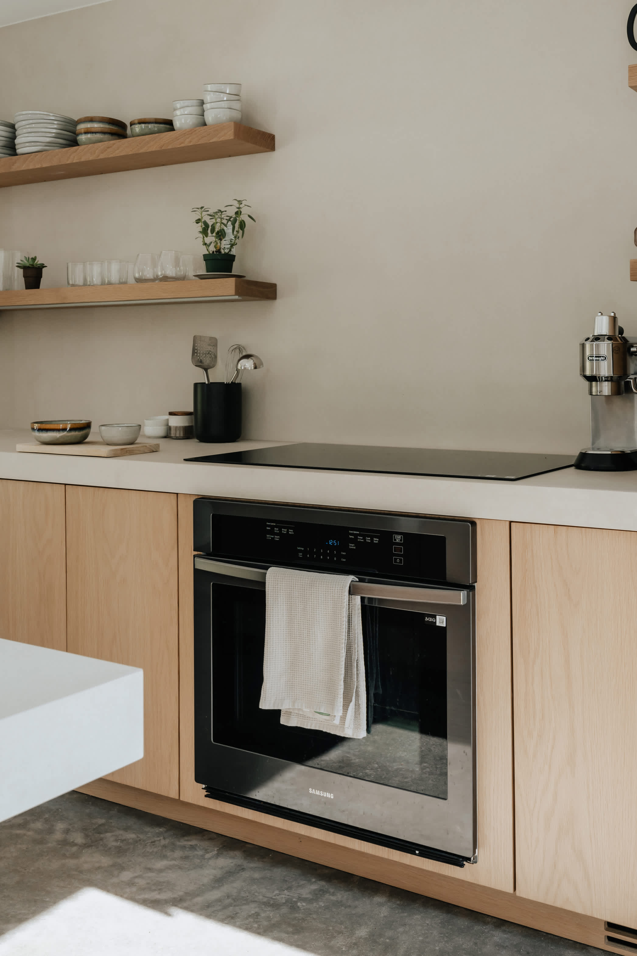 A close-up of the modern kitchen showcasing light wood flat-panel cabinetry, white countertops, and a built-in Samsung oven below an induction cooktop. Open wooden shelves display dishes and plants.