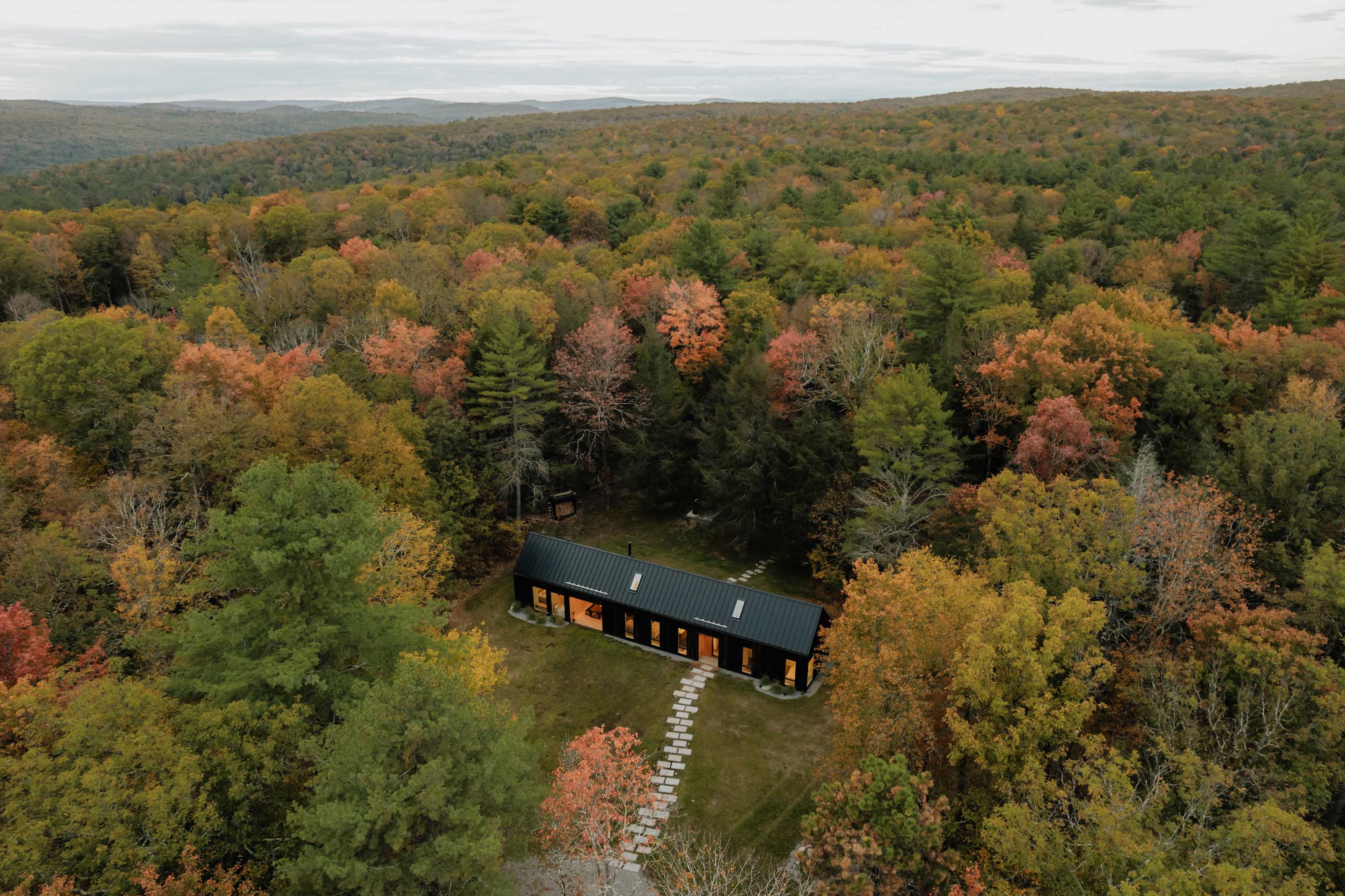 Aerial view of the dark, modern house nestled in a vast, vibrant autumn forest, showcasing its secluded location and the colorful fall foliage.
