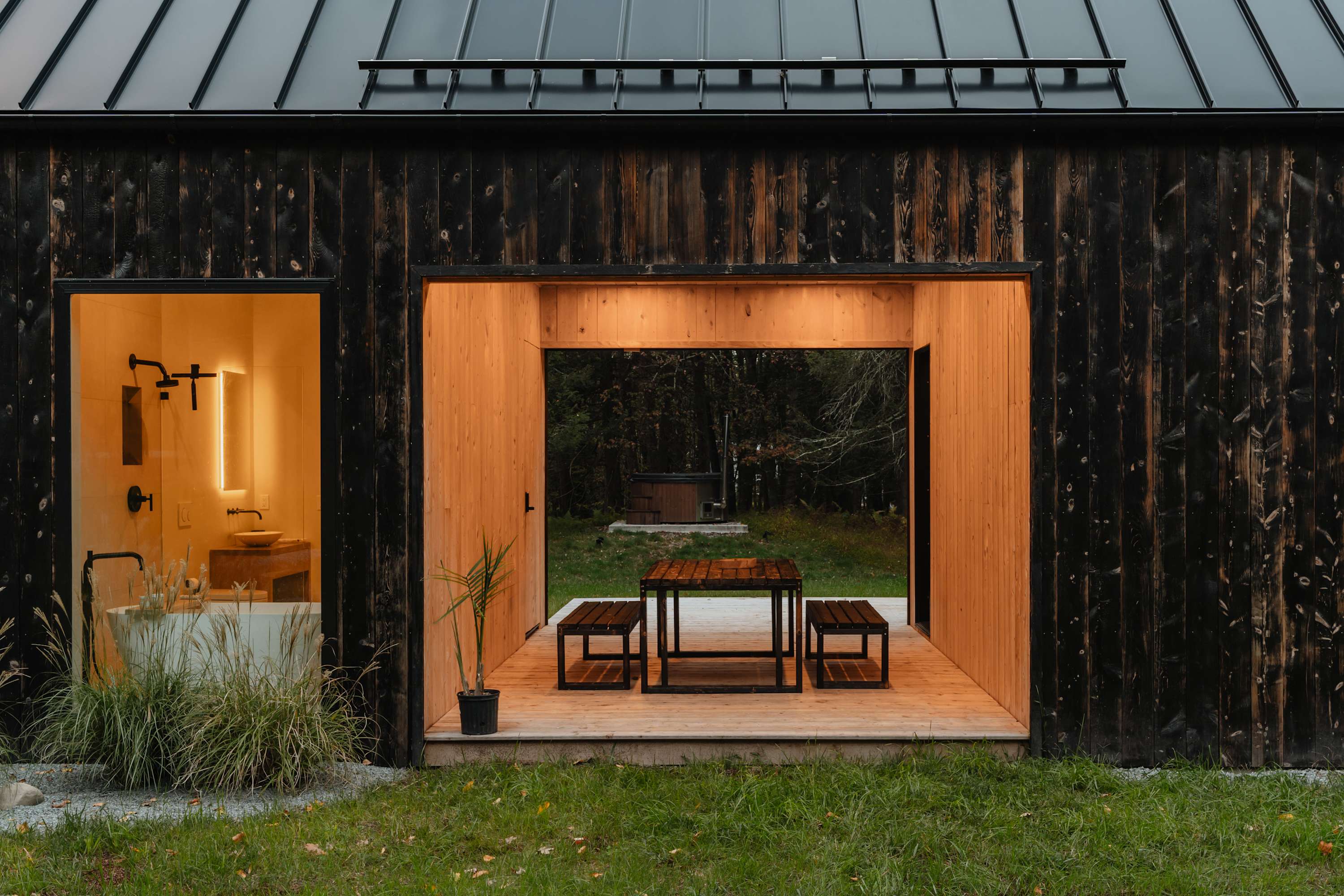 The back of a modern black-stained wood cabin features a large open-air covered patio with a dining set, leading to a grassy yard and a hot tub in the distance. A brightly lit bathroom with a freestanding tub is visible to the left.