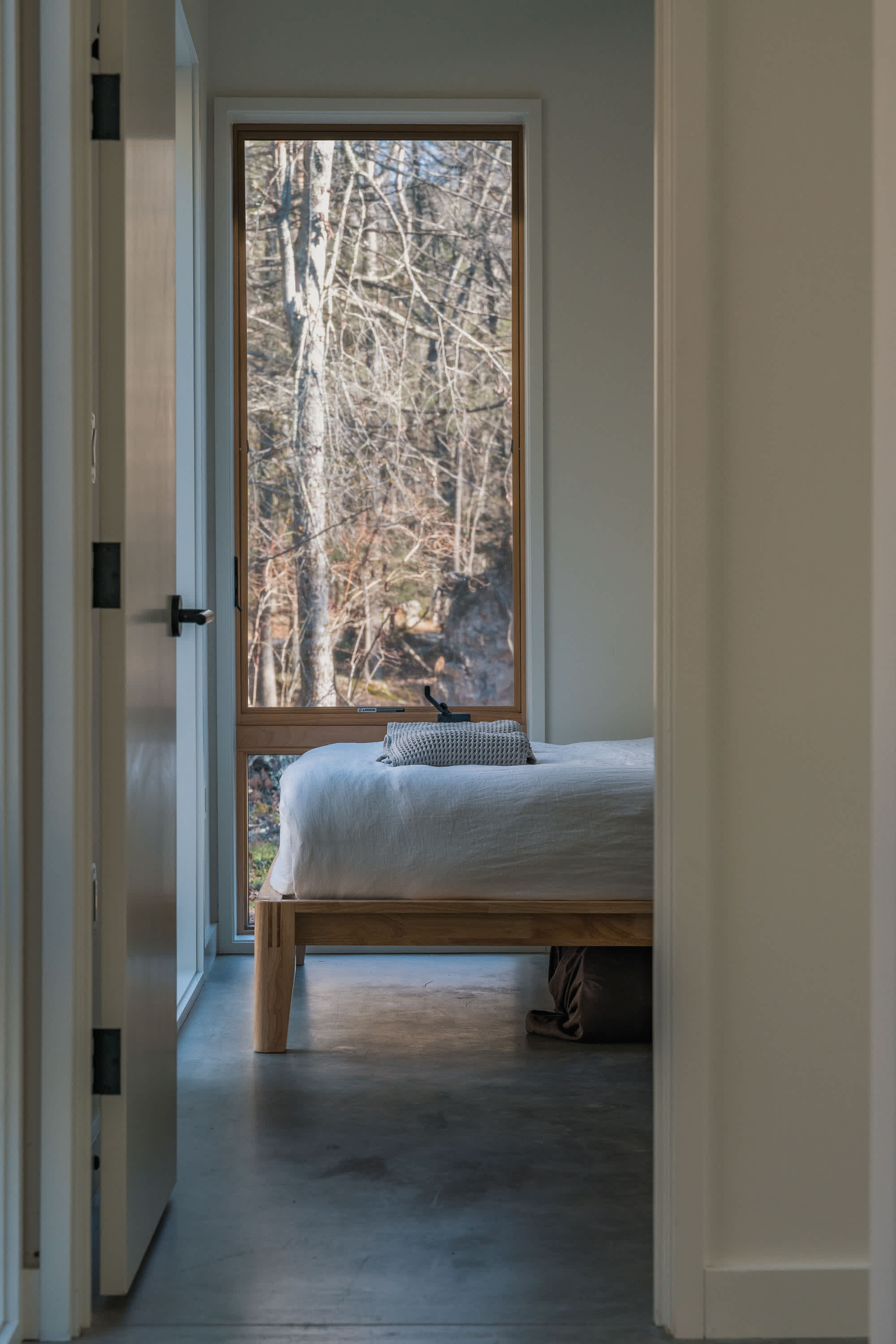A view from a hallway looking into a minimalist bedroom with a low-profile wooden bed and a large window showcasing a winter forest landscape. The room maintains a clean, bright aesthetic with concrete floors.