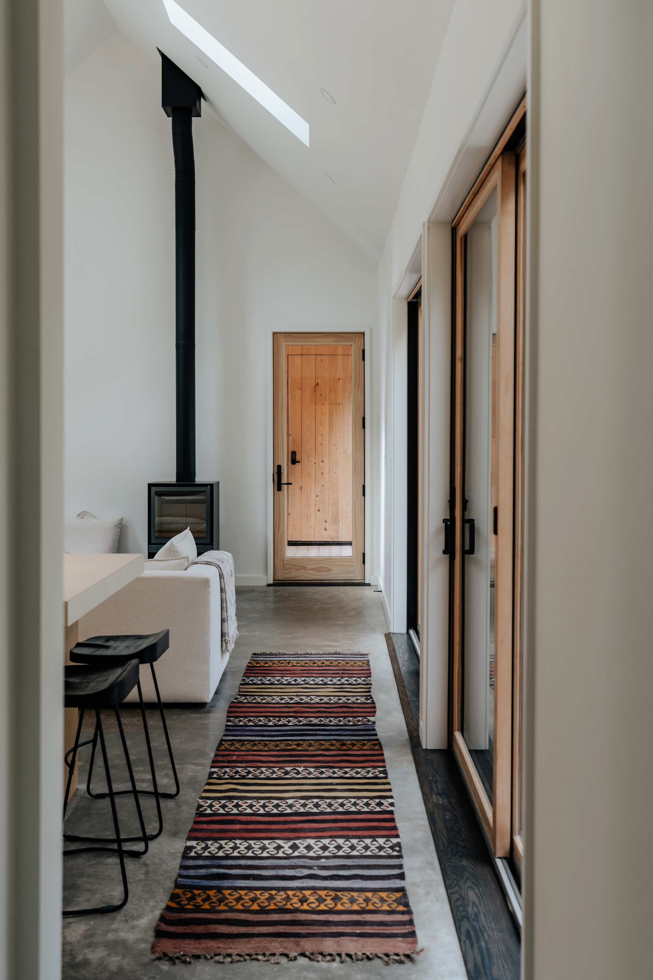 Bright hallway leading to a wooden front door, featuring a colorful striped rug, a glimpse of the living area with a wood stove, and a large window.