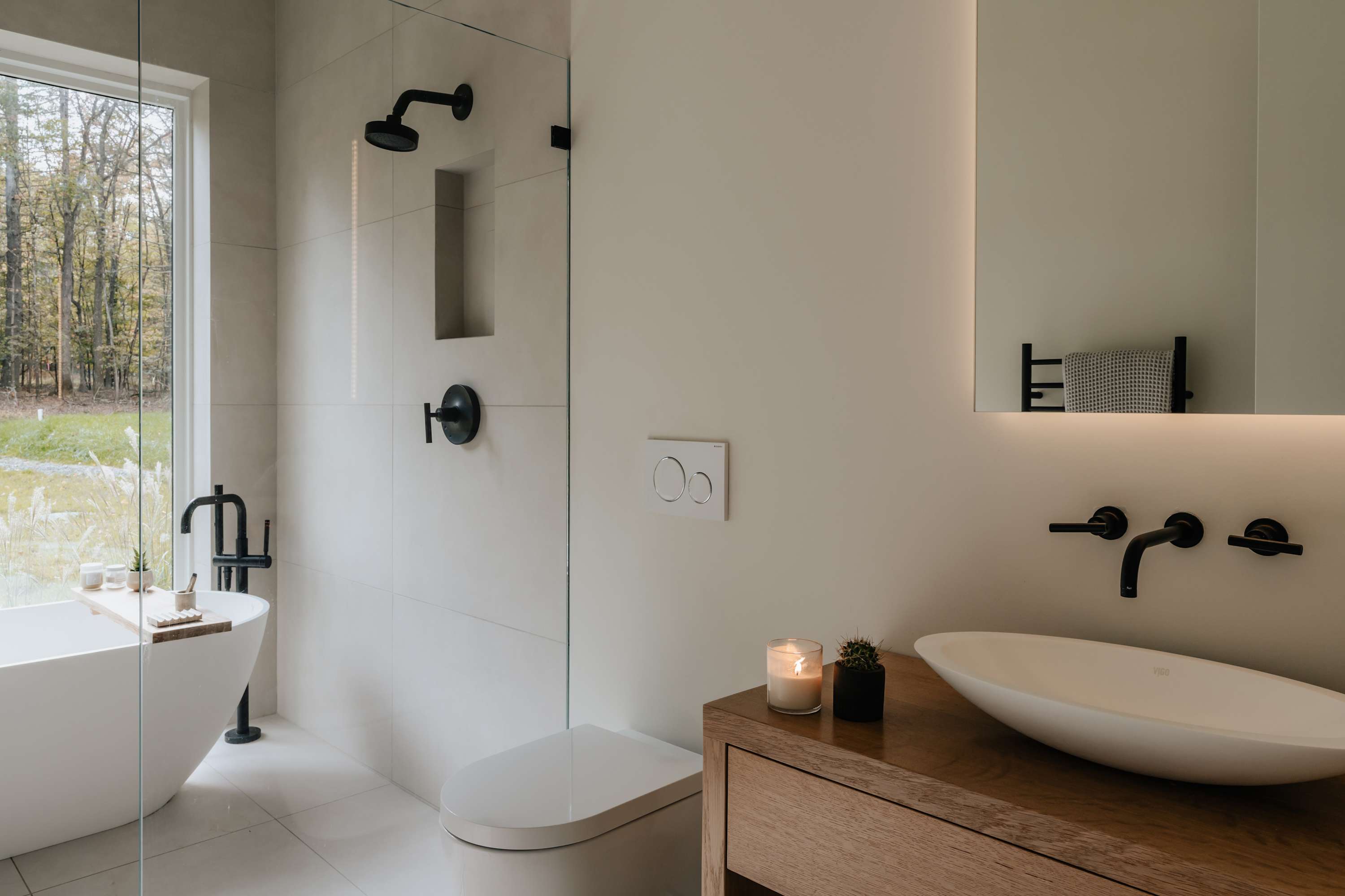 Modern bathroom with light-colored tiled walls, a glass-enclosed shower, a wall-mounted toilet, and a vessel sink on a wooden vanity, with a freestanding tub visible near a large window.