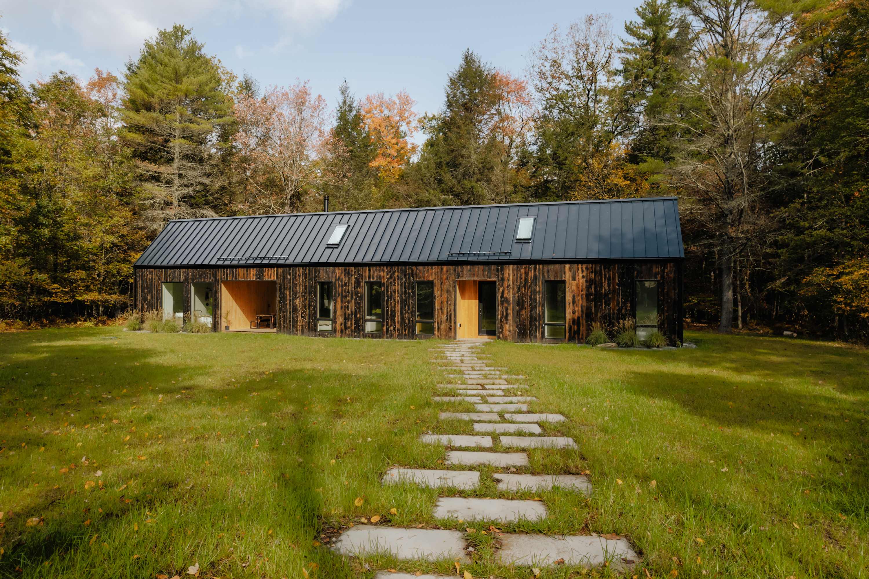 A long, dark-clad modern house with a black metal roof and skylights, viewed from a grassy lawn with a stone slab pathway leading towards the main entrance. The house is surrounded by a dense forest with autumn colors, creating a picturesque and inviting approach.