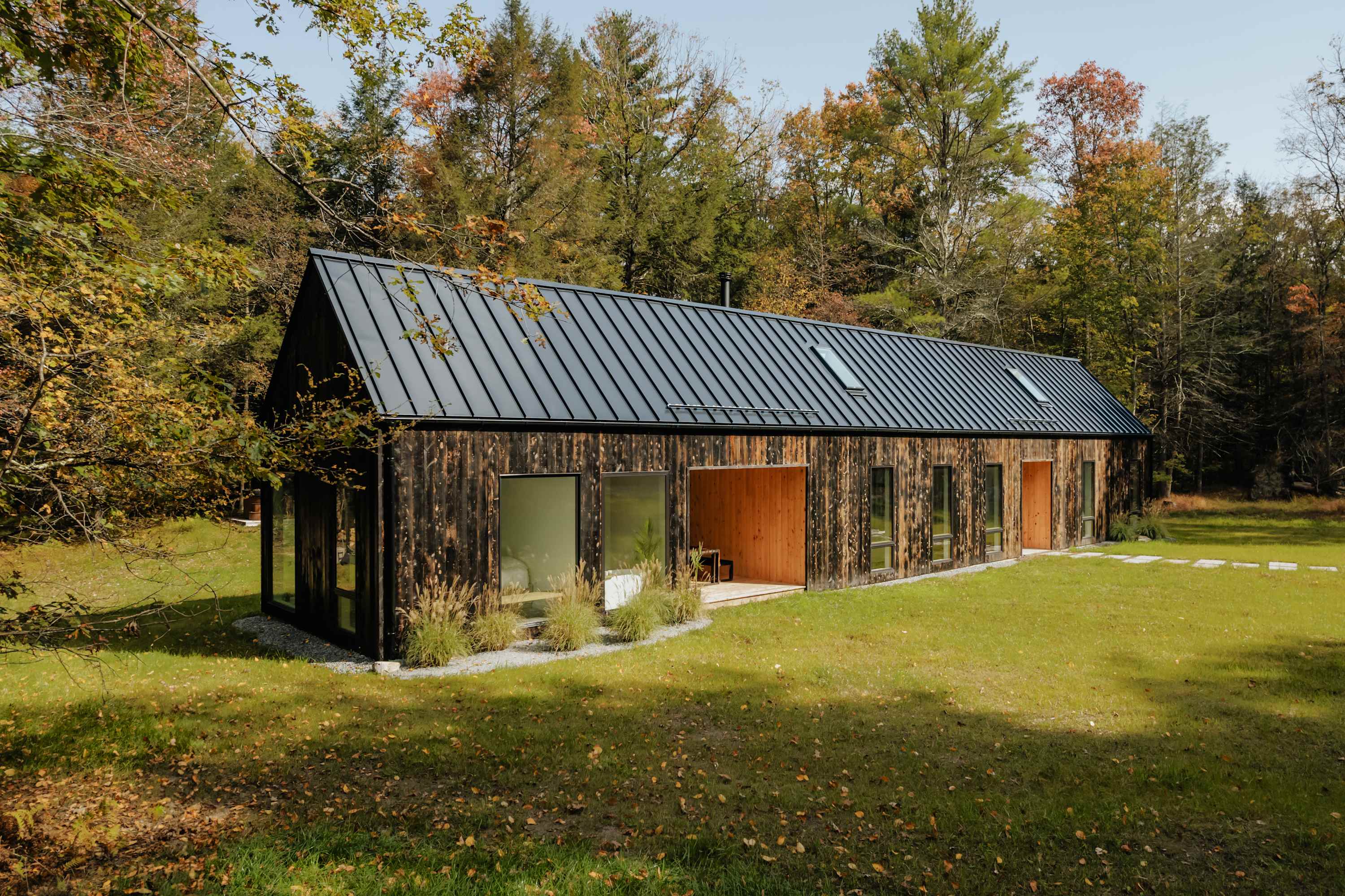 A long, modern dark-clad house with a black metal roof and multiple skylights, set in a clearing surrounded by a vibrant autumn forest. The house features large windows and a recessed wooden entrance, showcasing contemporary design in a natural setting.
