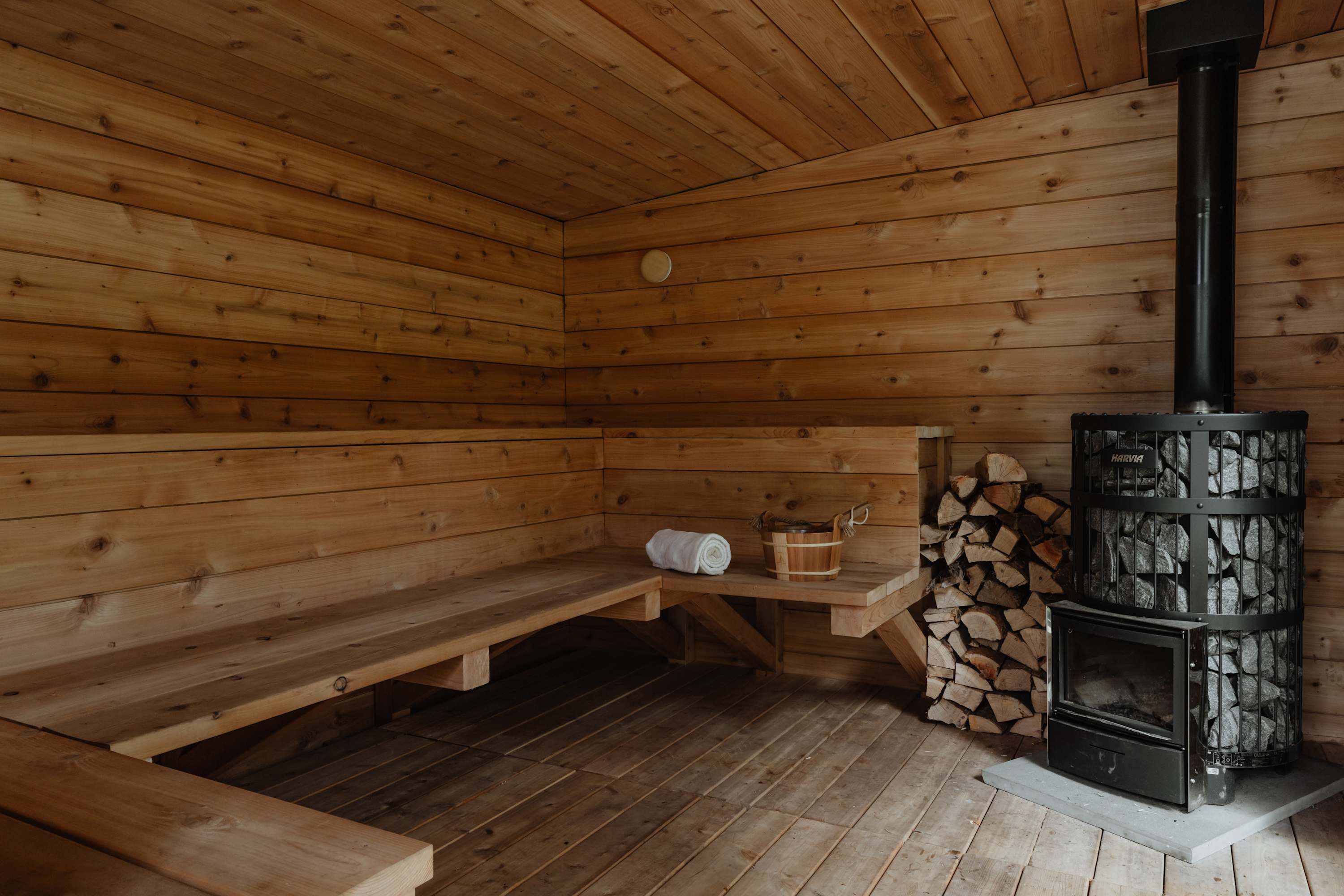Rustic wooden sauna interior with slatted benches, a prominent wood-burning stove with a stone-filled top, and a bucket and ladle, ready for a relaxing experience.