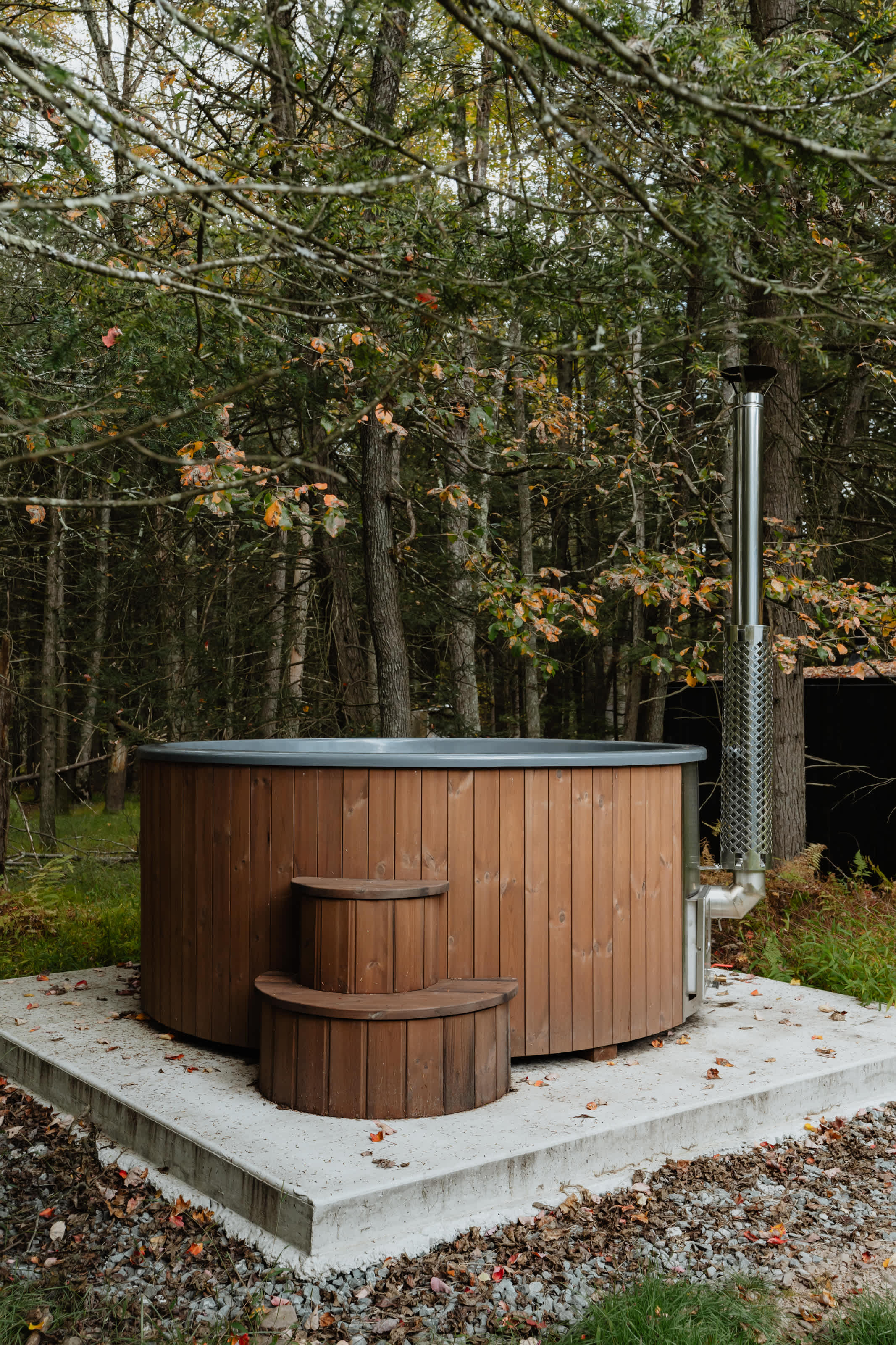 A round, wooden hot tub with a metal chimney for heating, set on a concrete pad in a lush, wooded area with autumn foliage. Wooden steps lead up to the tub, blending with the natural surroundings.