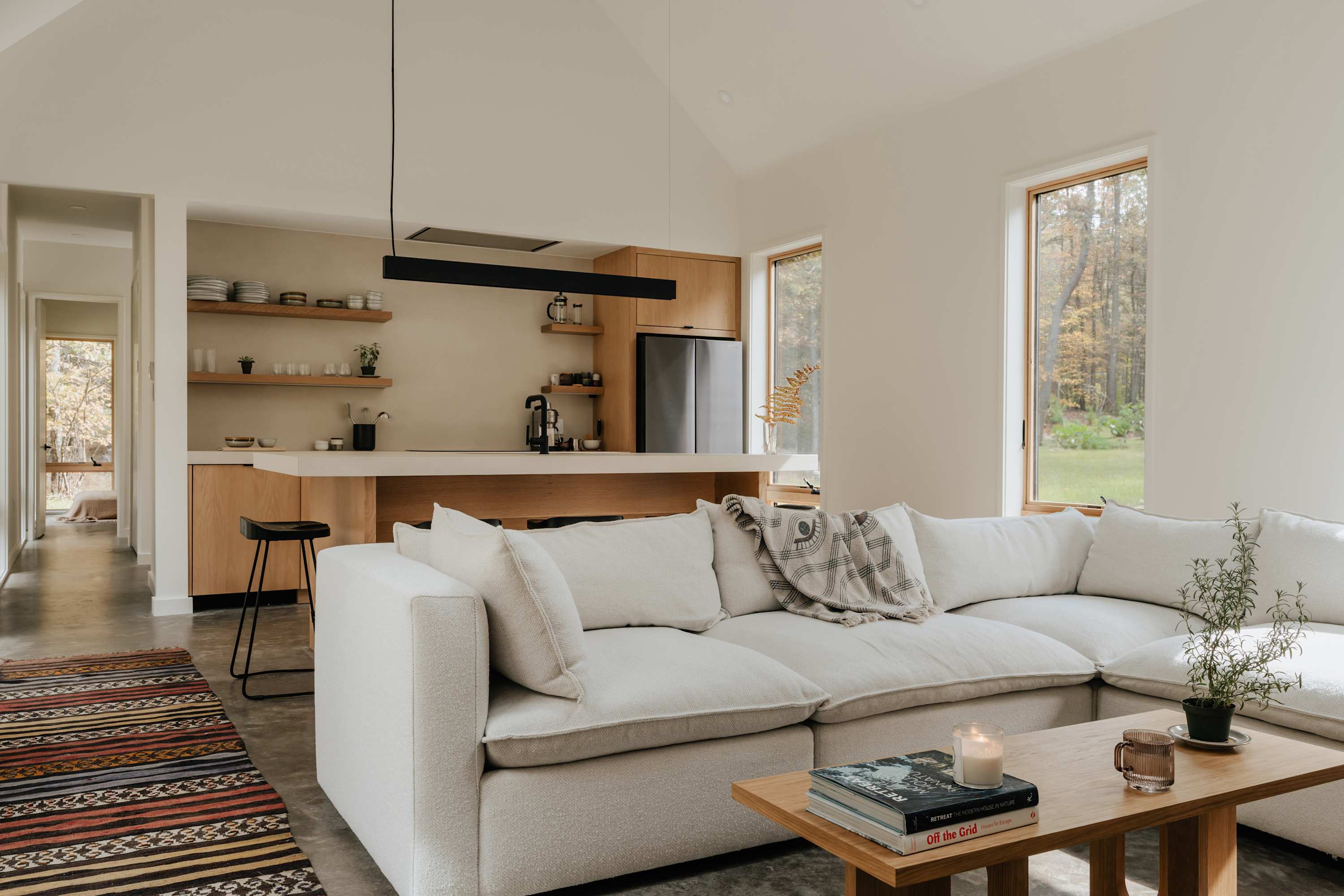 Open-concept living room and kitchen with a large white sectional sofa, a wooden coffee table, and a colorful rug, offering views of the surrounding forest through large windows.