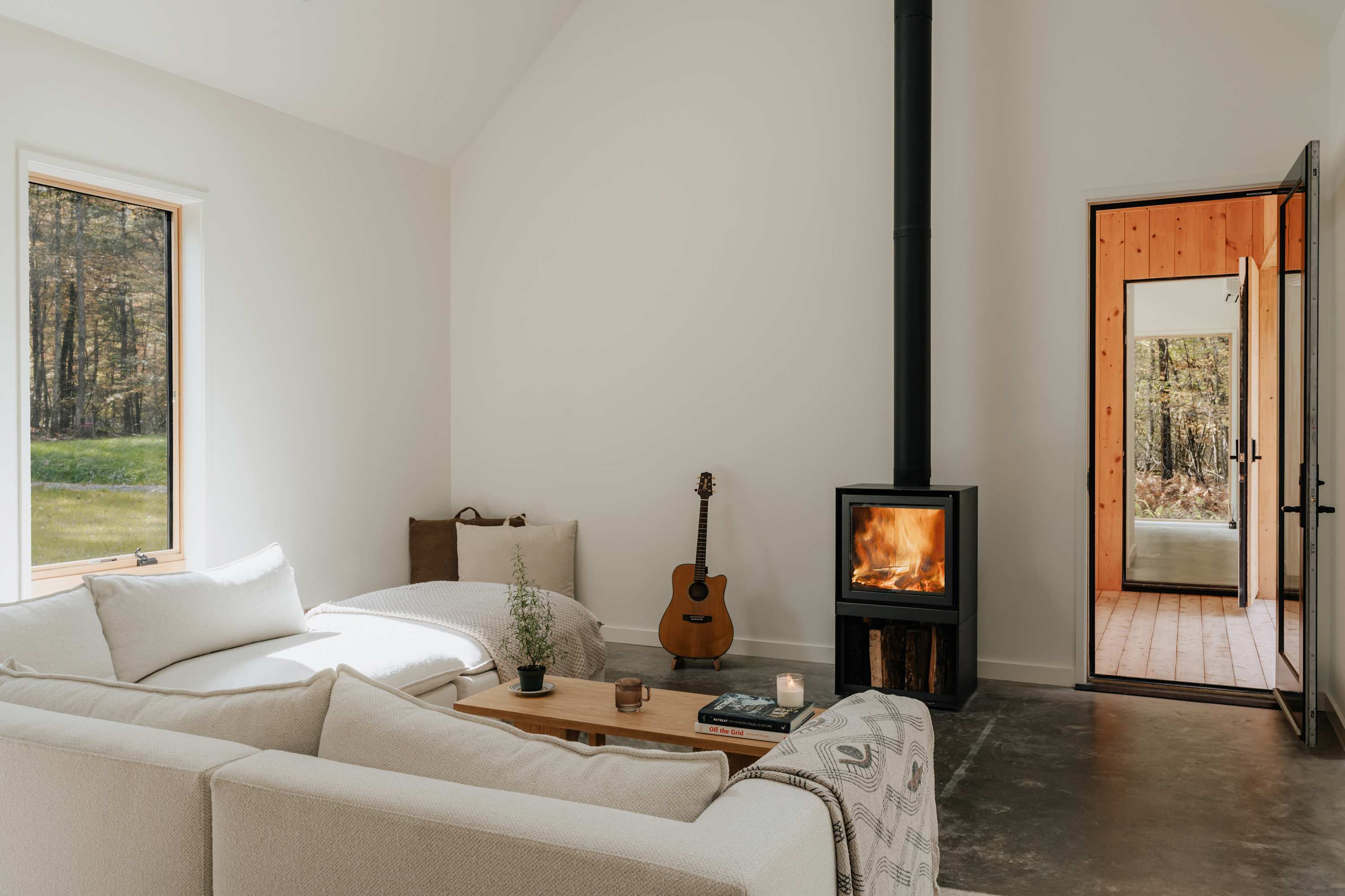 A bright, minimalist living room features a light-colored sectional sofa, a modern black wood-burning stove with a visible flame, and a wooden coffee table. Large windows offer views of the surrounding forest, and a guitar leans against the wall.