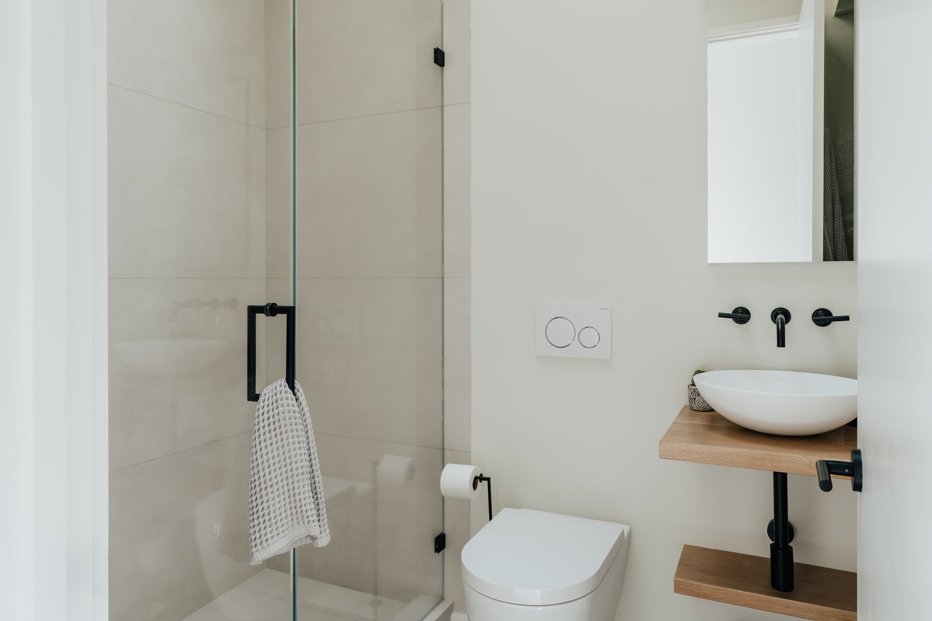 Modern minimalist bathroom with light-colored tiled walls, a glass-enclosed shower, a wall-mounted toilet, and a vessel sink on a floating wooden shelf with black fixtures.