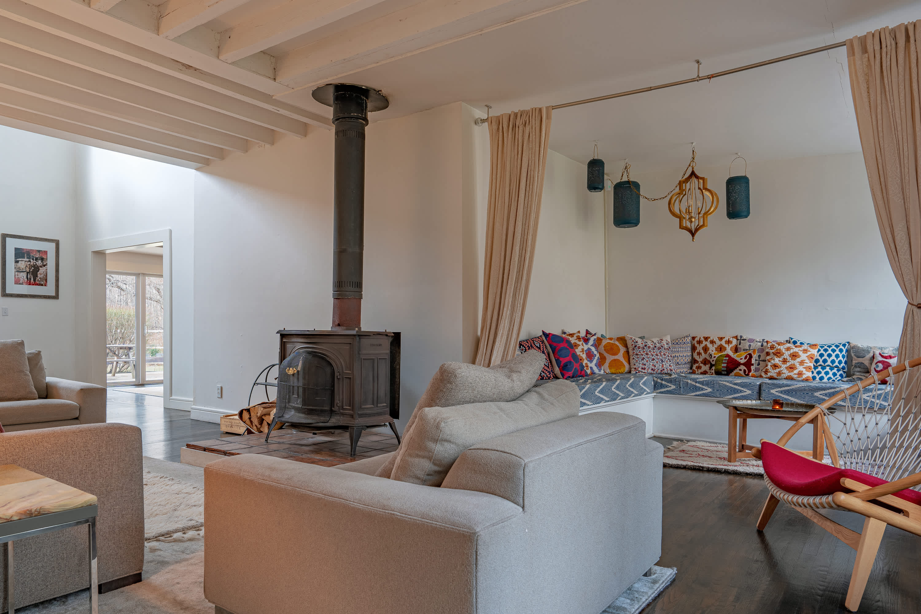 A cozy living room features a prominent black wood-burning stove on a tiled hearth, surrounded by light-colored sofas and a built-in bench seating area with colorful pillows and draped curtains, under exposed white beams.
