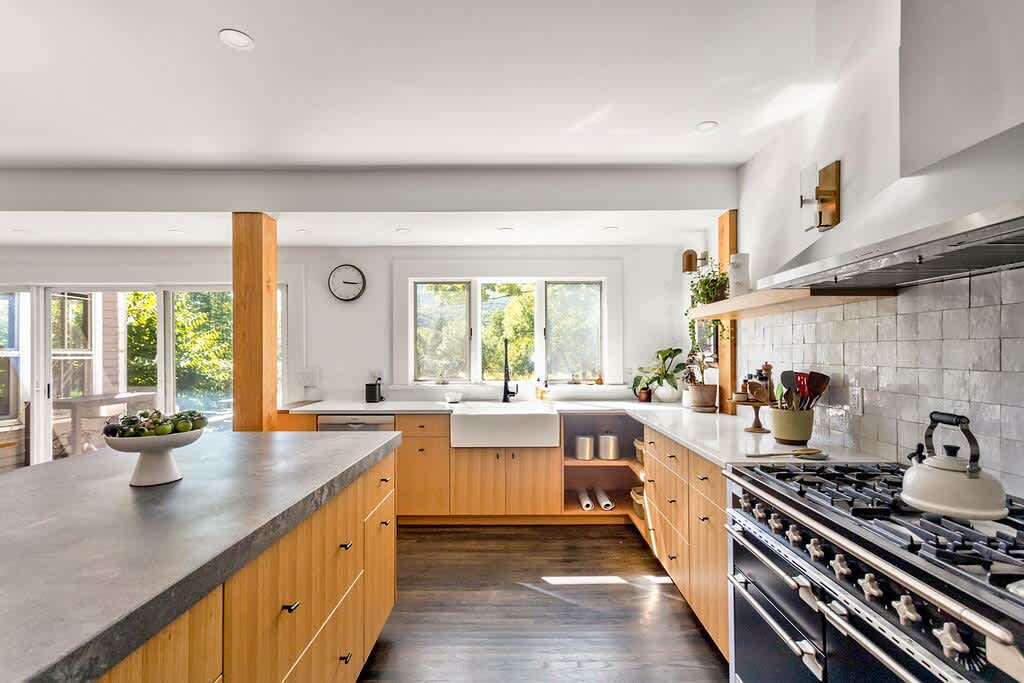 A spacious, modern kitchen boasts light wood cabinetry, white countertops, a large kitchen island with a concrete-like top, and a professional-style range with a stainless steel hood, all under recessed lighting and dark hardwood floors.