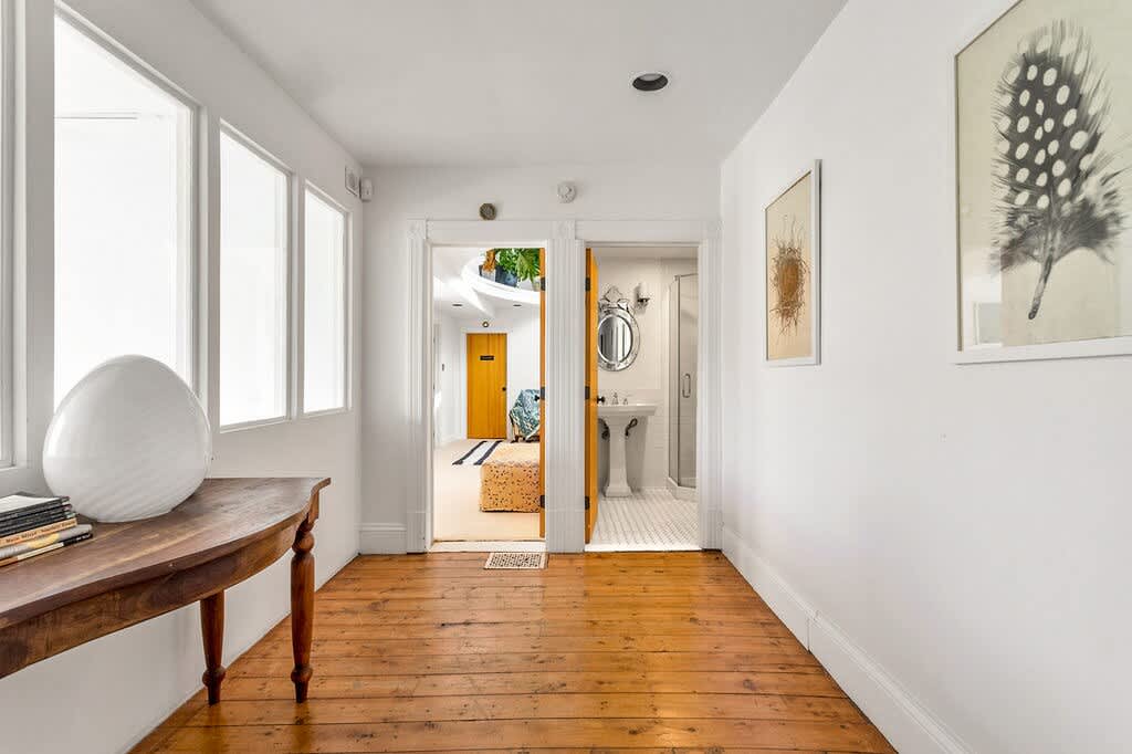Sunny hallway with polished hardwood floors and white walls, featuring a rustic wooden console table with a large white decorative egg. Two open doorways lead to other rooms, one revealing a bedroom and the other a bathroom with a pedestal sink.