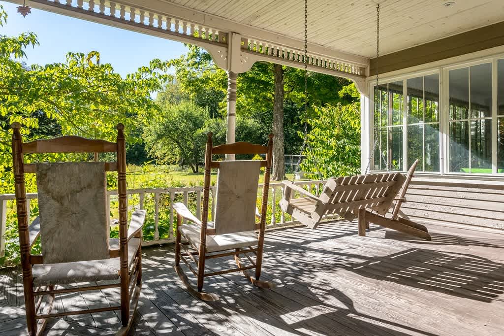 A sunny wooden porch features two classic rocking chairs with light fabric cushions and a slatted wooden porch swing, overlooking a lush green garden and trees.