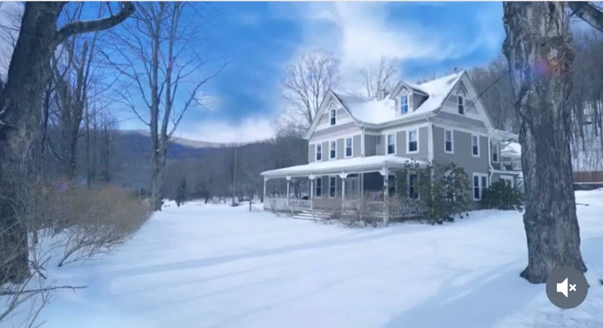 Picturesque two-story house with gray siding and a wide porch, nestled in a snow-covered landscape with bare trees and distant mountains under a bright blue sky.