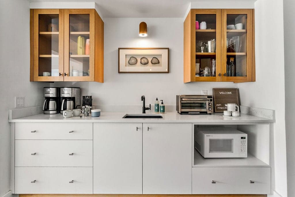 Compact kitchenette with white shaker-style base cabinets, a white countertop, a sink, and two light wood upper cabinets with glass fronts, featuring a microwave and coffee makers.