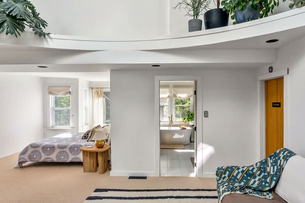 Bright and airy bedroom area with a bed featuring a patterned quilt, light wood side table, and a view of the outdoors through large windows, connected to a separate bathroom area visible through a doorway.