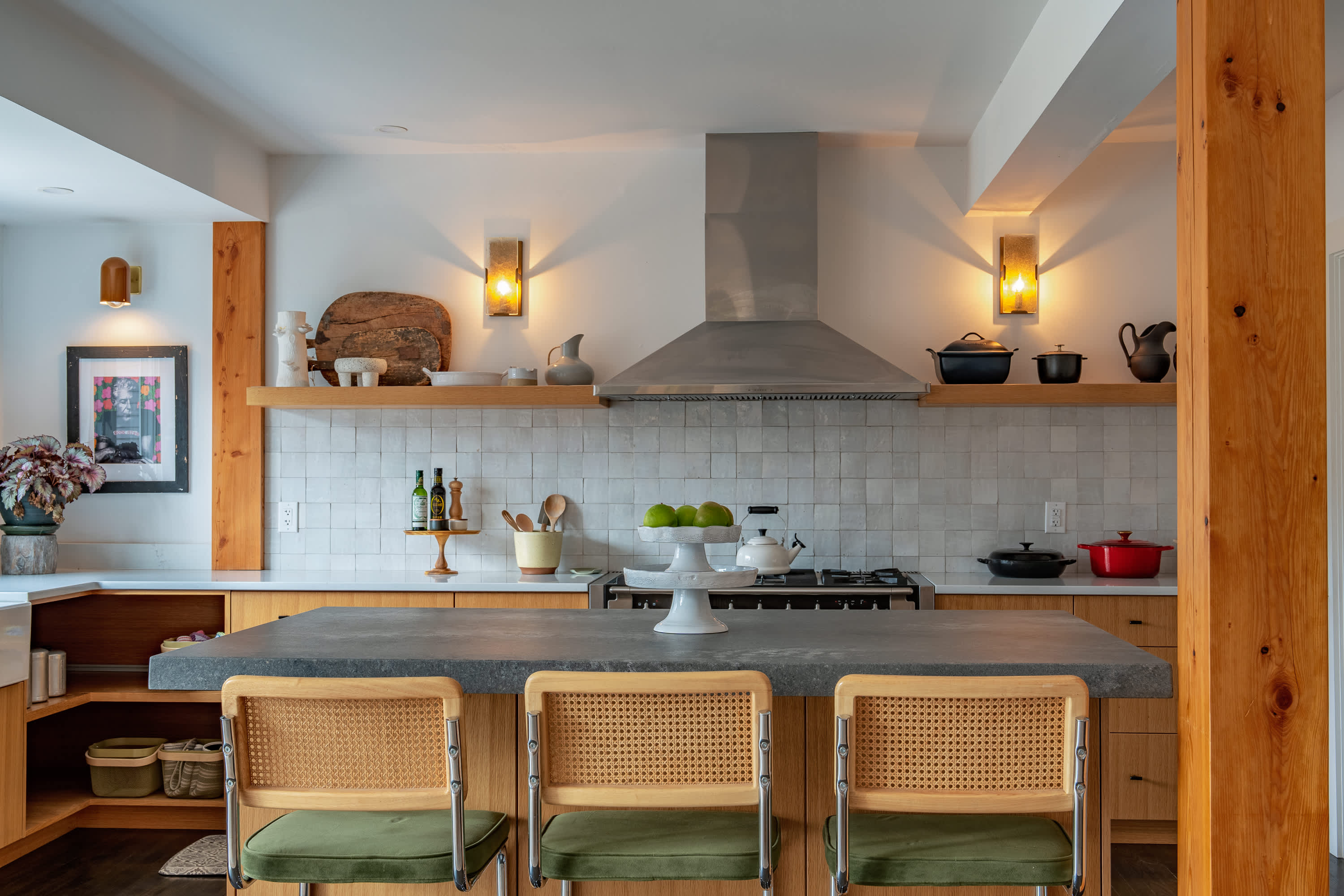 Modern kitchen featuring a large concrete island with cane-backed bar stools, light wood cabinetry, a stainless steel range hood, and white square tile backsplash, illuminated by two wall sconces.