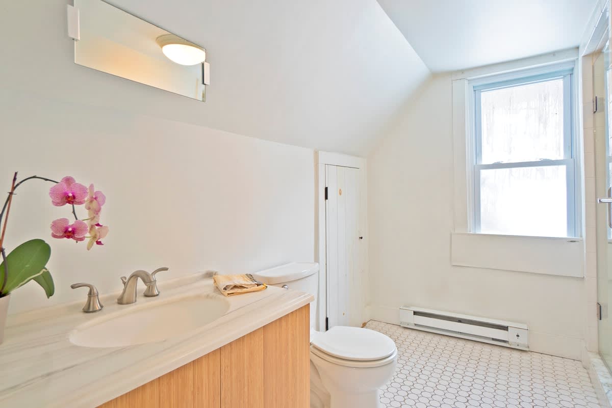 Bright bathroom with white walls, a light wood vanity with a marble countertop, a white toilet, and a window with frosted glass, featuring a hexagonal tile floor.