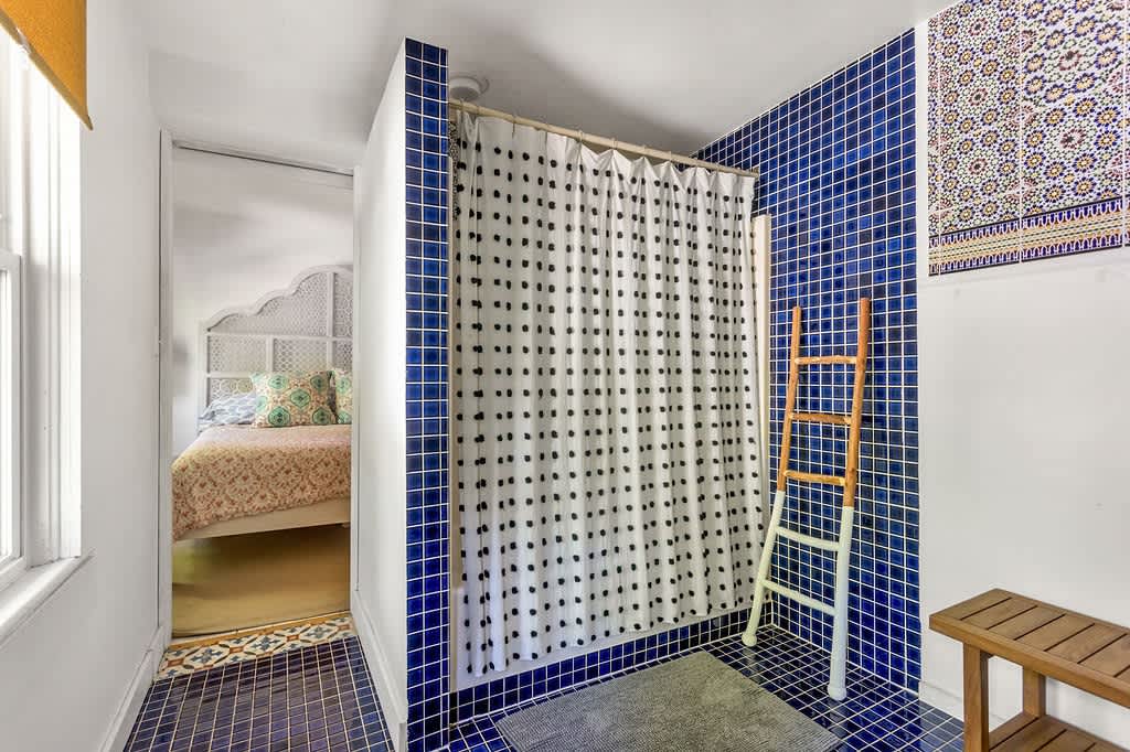 Vibrant bathroom featuring dark blue square tiles on the walls and floor, surrounding a shower with a white and black polka dot curtain. A wooden ladder acts as a towel rack, and a glimpse of a bedroom with a white headboard is visible through the doorway.