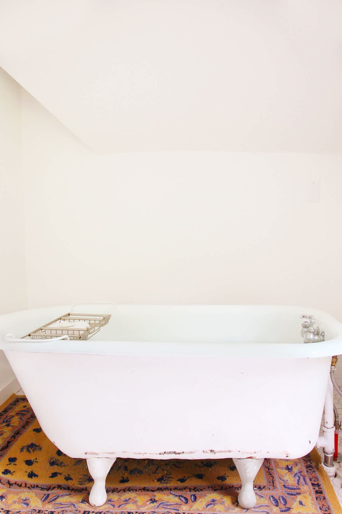 Minimalist bathroom featuring a white clawfoot bathtub with a metal caddy, placed on a colorful patterned rug, against plain white walls.
