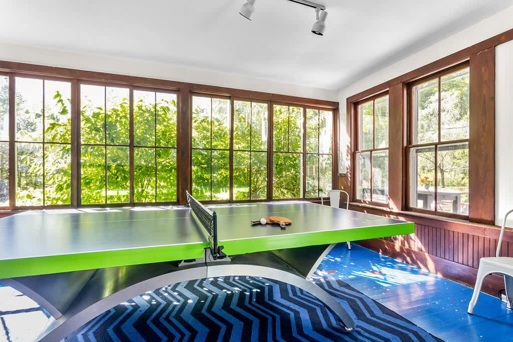 Bright game room with a vibrant green and black ping-pong table, set on a blue patterned rug, surrounded by large windows offering views of lush greenery, creating an inviting recreational space.