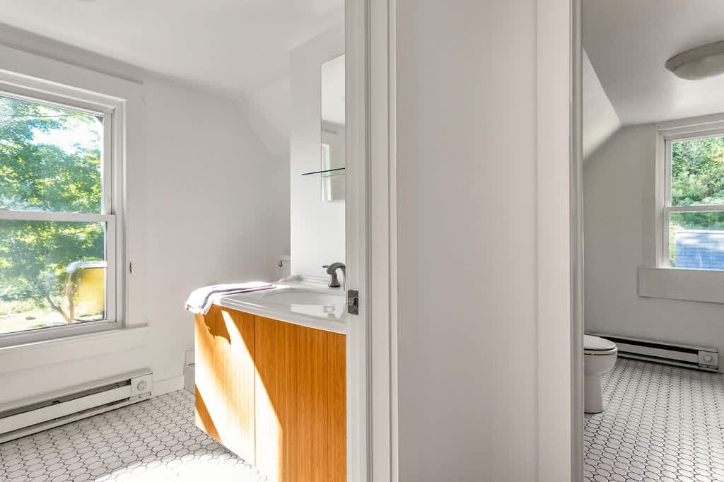 Bright bathroom with white hexagonal tile flooring and a light wood vanity with a white sink, positioned under a large window. A mirror is visible above the sink, and a toilet is partially seen in an adjacent alcove.