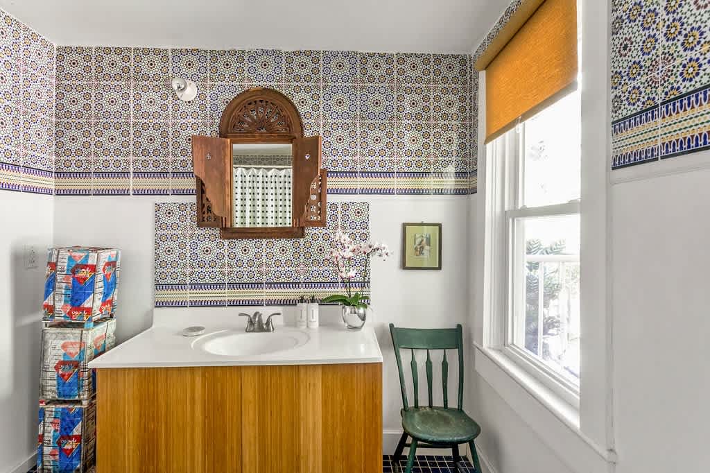 Bright bathroom with white walls and a striking patterned tile backsplash featuring blue, yellow, and brown geometric designs. A wooden vanity with a white countertop and sink is topped by an ornate carved wooden mirror cabinet, beside a window with a brown blind.