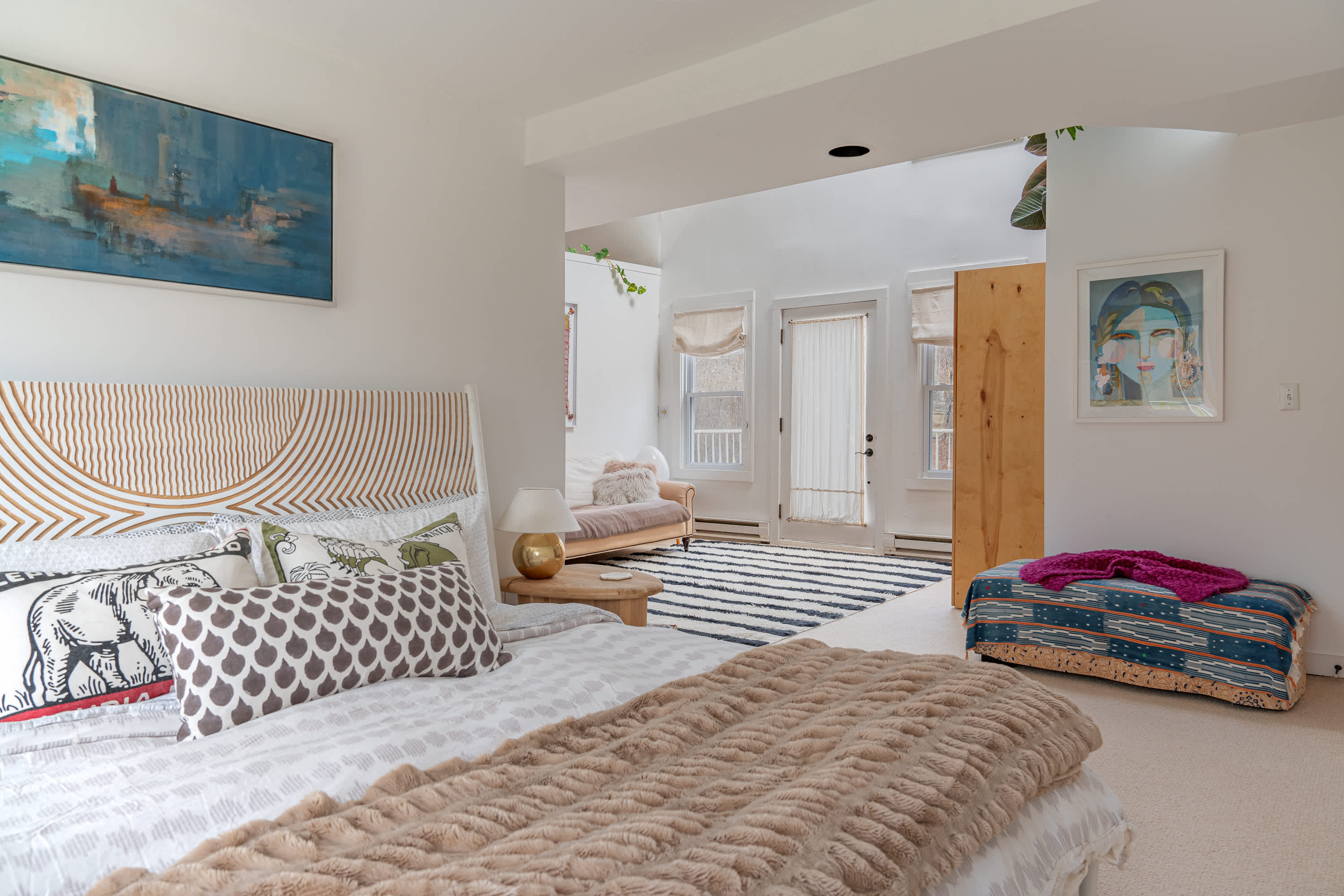 Expansive master suite featuring a queen bed with a textured headboard, a plush throw, and a large abstract painting, leading into a sitting area with a striped rug and access to an outdoor space.