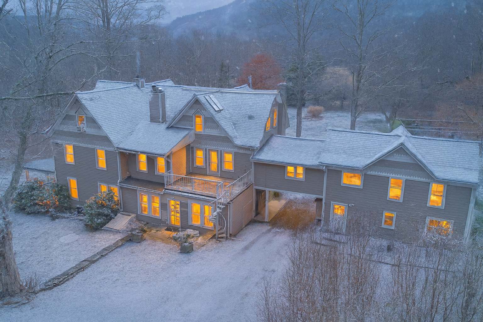 Aerial view of a large, sprawling house with multiple wings and gables, covered in a light dusting of snow, with warm lights glowing from numerous windows, set against a backdrop of snow-covered hills and trees.