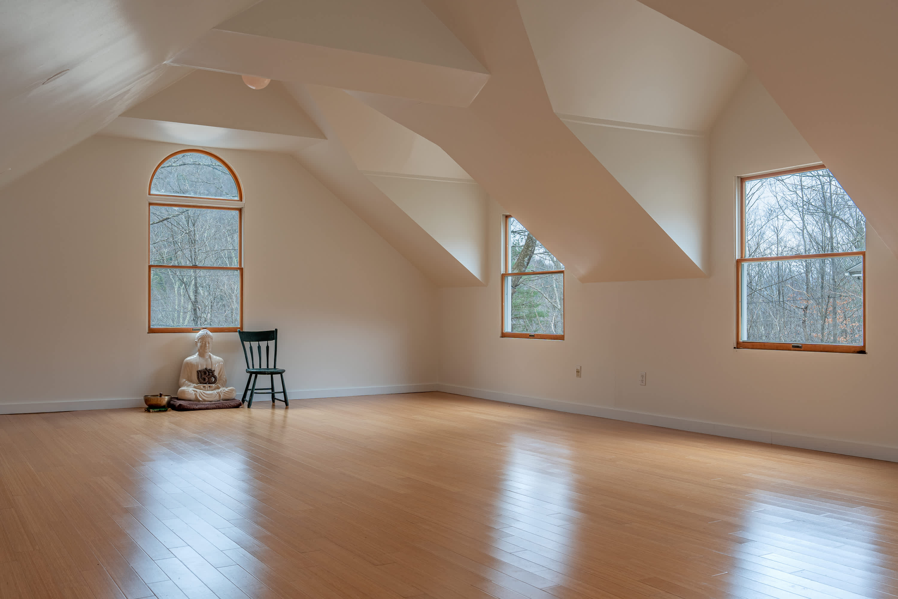 Spacious attic yoga studio with light hardwood floors, sloped ceilings, and multiple windows offering views of the surrounding trees, featuring a Buddha statue and a meditation bowl.
