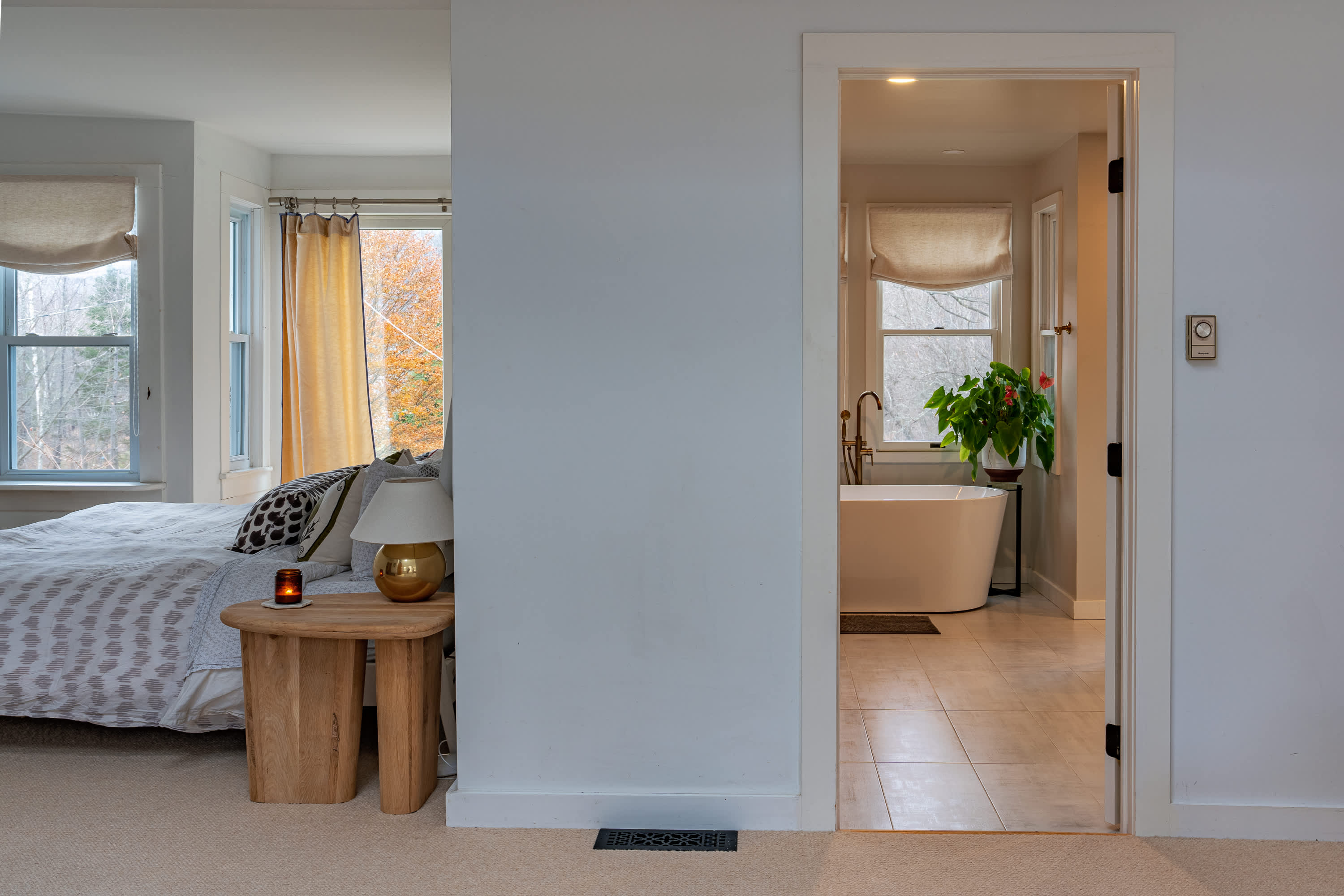 View from a bedroom into an adjacent spacious bathroom, revealing a freestanding white bathtub and large windows, with a light wood side table and lamp in the foreground.