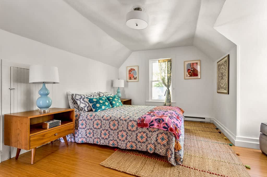 Cozy bedroom with a patterned bedspread, two wooden nightstands with blue lamps, and a window with patterned curtains, set on a light wood floor with a woven rug.