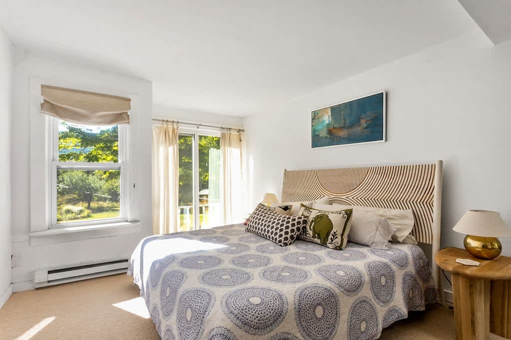 Cozy bedroom with a queen-sized bed featuring a unique carved wooden headboard and a blue and white patterned quilt, positioned next to a large window offering views of lush greenery.