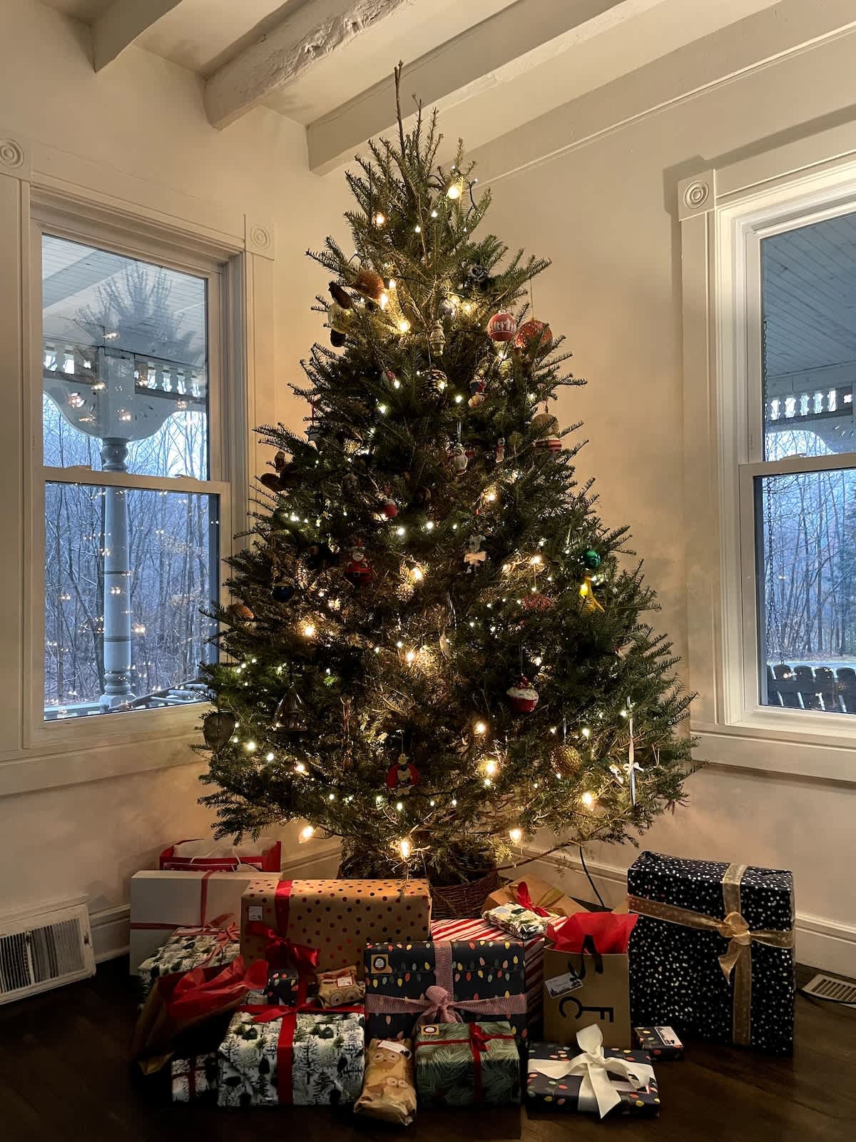 A festive Christmas tree, brightly lit with warm white lights and adorned with colorful ornaments, stands in a cozy room surrounded by numerous wrapped presents, with windows revealing a winter landscape outside.