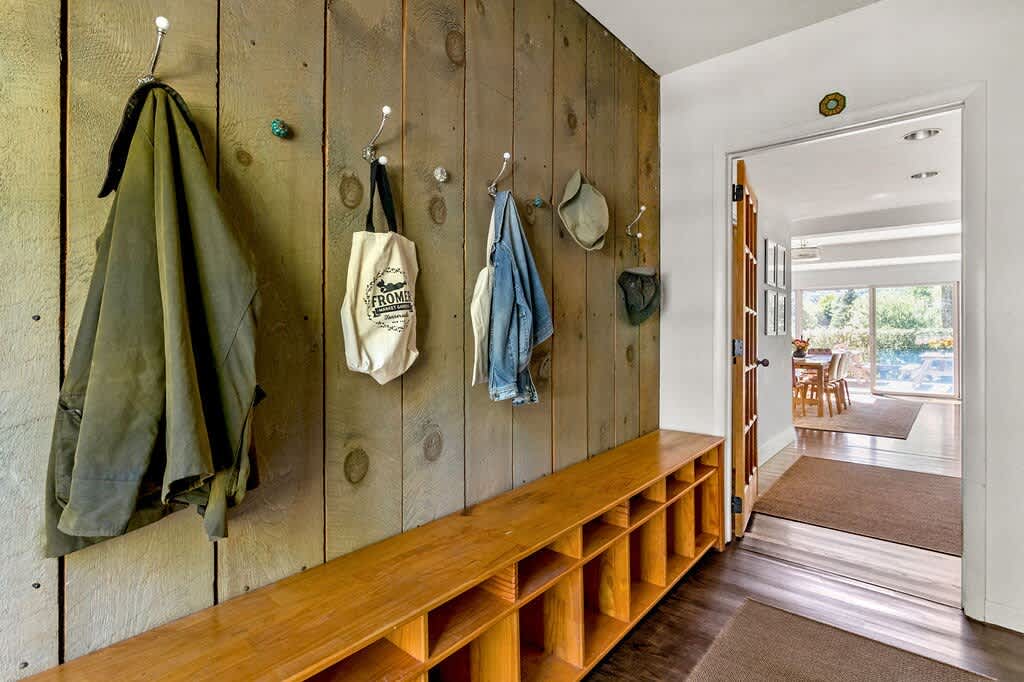 A rustic mudroom features a wall clad in vertical wooden planks with multiple coat hooks holding jackets and bags, above a long, built-in wooden bench with cubby storage, and dark hardwood floors.
