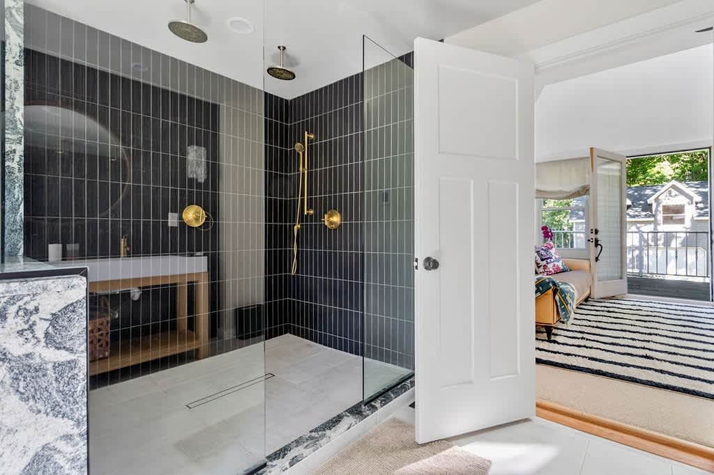 Modern walk-in shower with dark vertical subway tiles and a glass enclosure, featuring dual rain shower heads and gold fixtures, connected to the master bedroom suite.