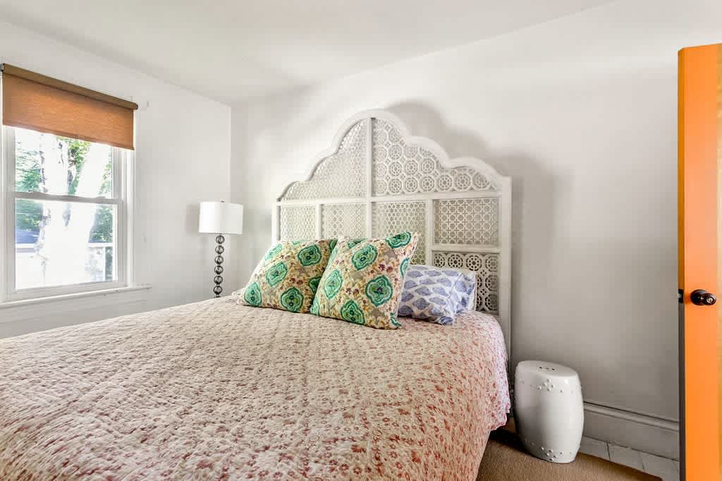 Bright bedroom with white walls and a decorative white headboard, featuring a bed covered in a patterned quilt and two green and blue patterned pillows. A large window with a brown blind lets in natural light, and a white ceramic stool serves as a bedside table.