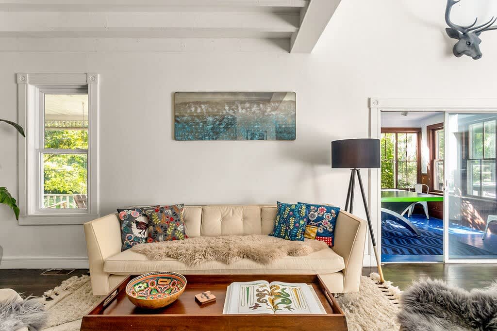 A bright living room showcases a light-colored sofa adorned with colorful pillows and a faux fur throw, positioned on a shaggy rug, with a wooden coffee table and a deer head wall decor, under exposed white beams.